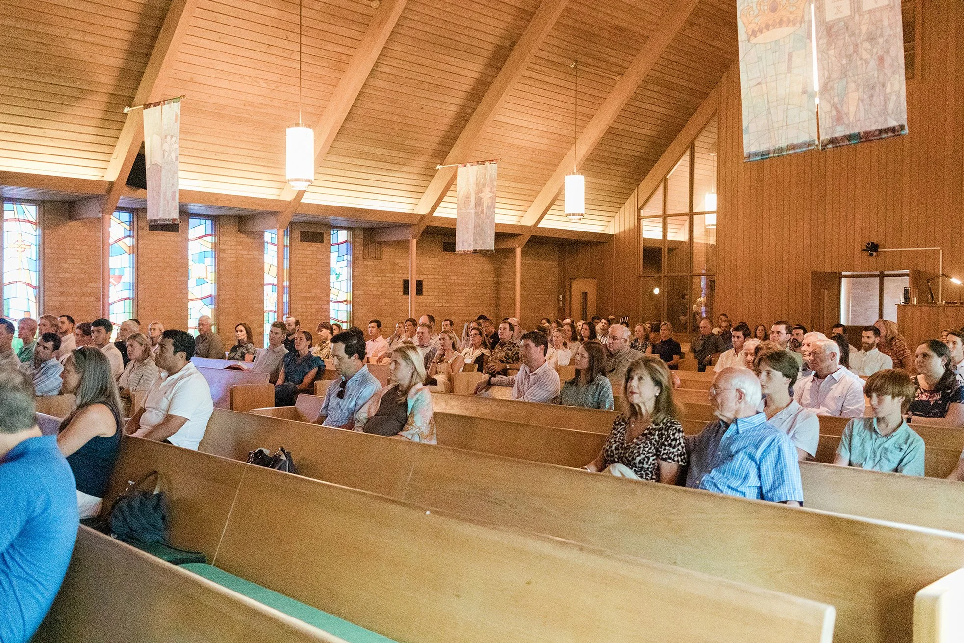 People seated in pews inside a church during a service or event, with wooden walls, stained glass windows, and hanging lights.