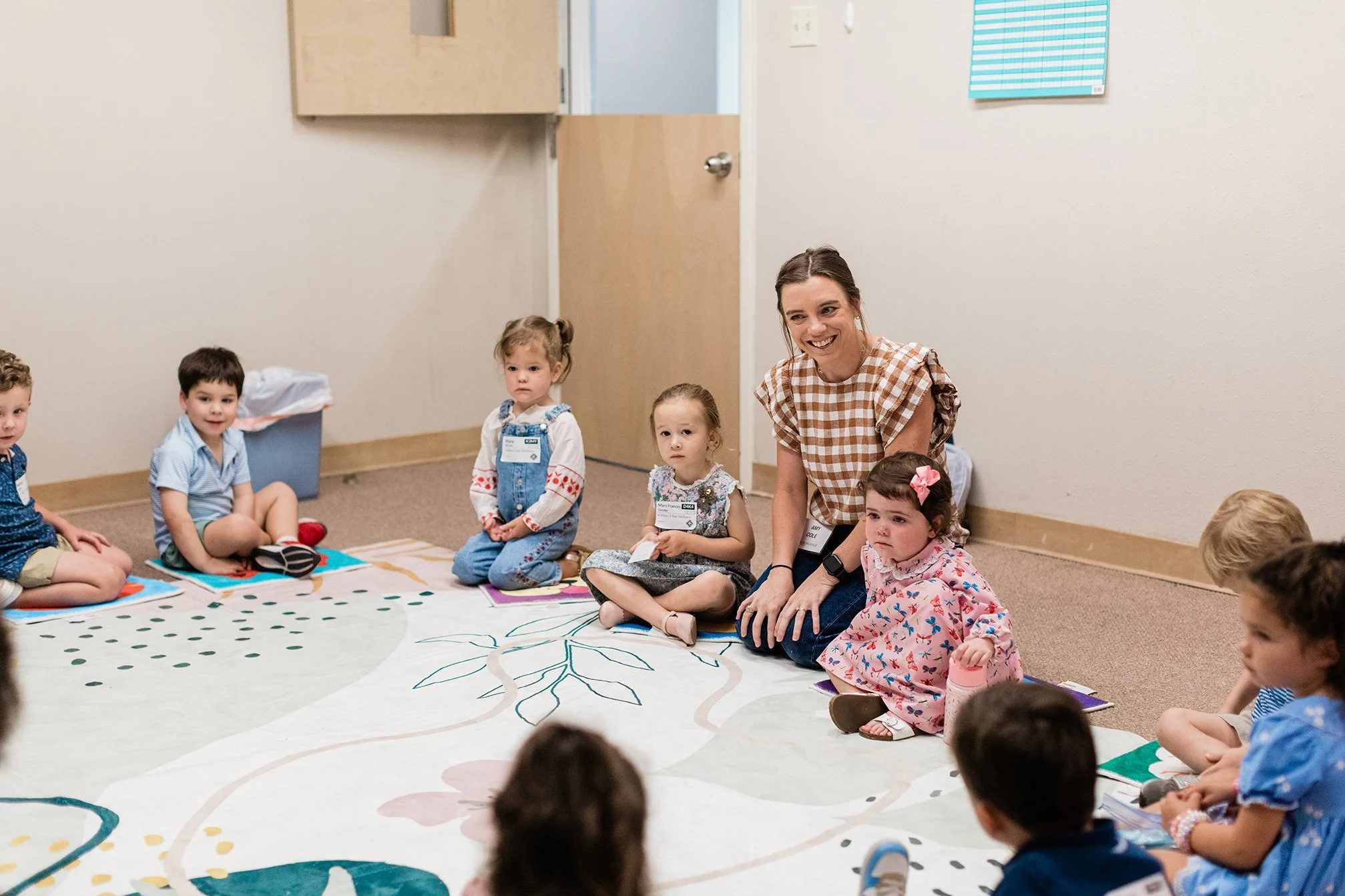 A teacher and five young children sitting on a large, colorful circular rug with a tree design in a classroom or daycare setting. The children are sitting cross-legged, some holding small objects and wearing name tags. The teacher, smiling, is kneeli