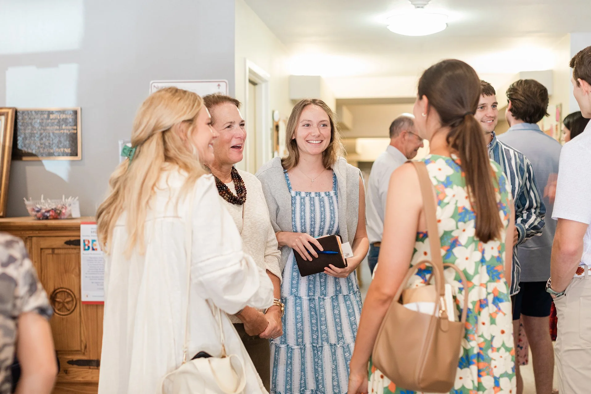 Group of people socializing indoors, including women in dresses and men in shirts, engaging in conversation and smiling at each other.