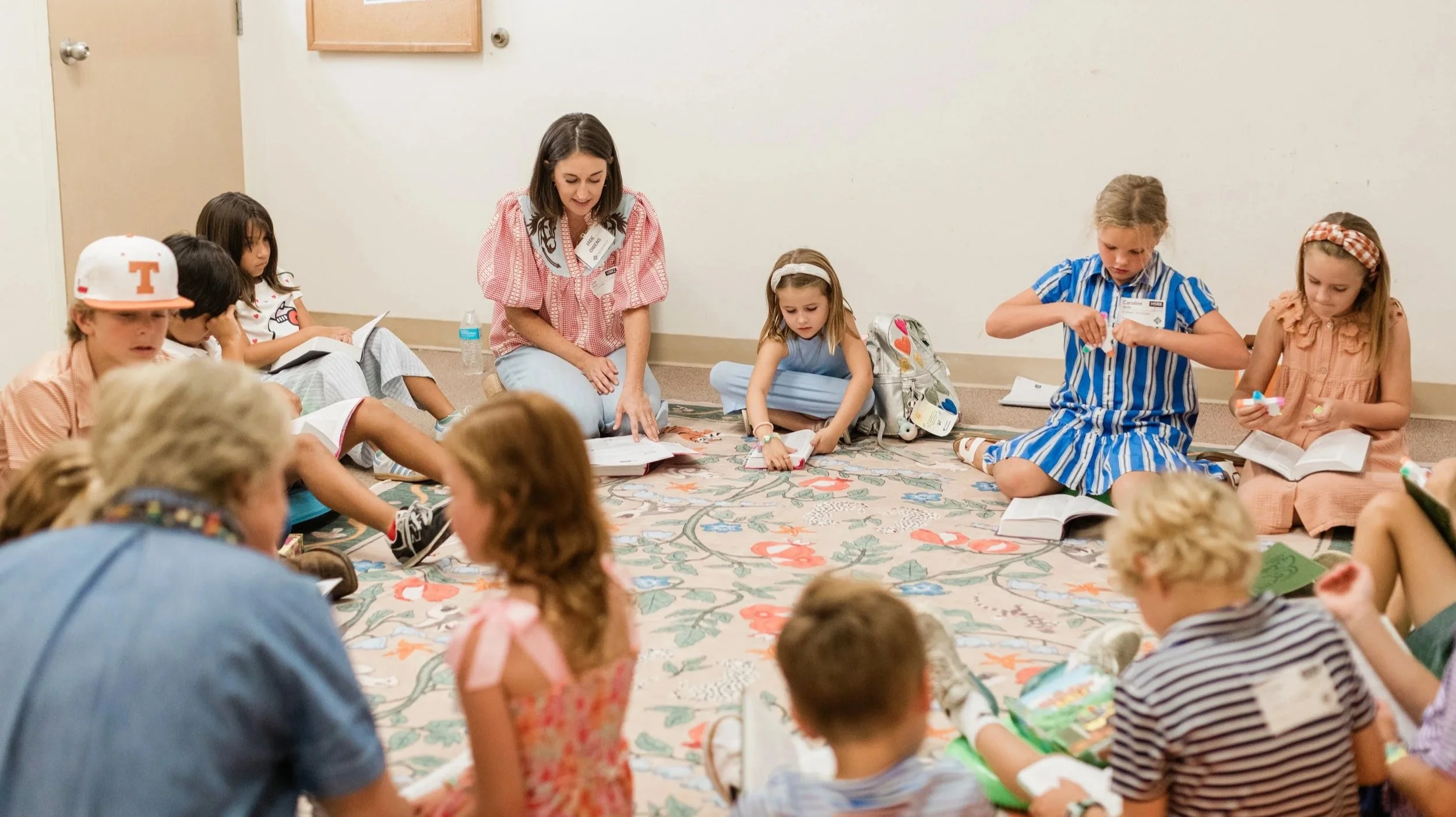 Children and a woman sitting on a carpeted floor in a circle, reading books and engaging in an activity, in a room with plain walls.