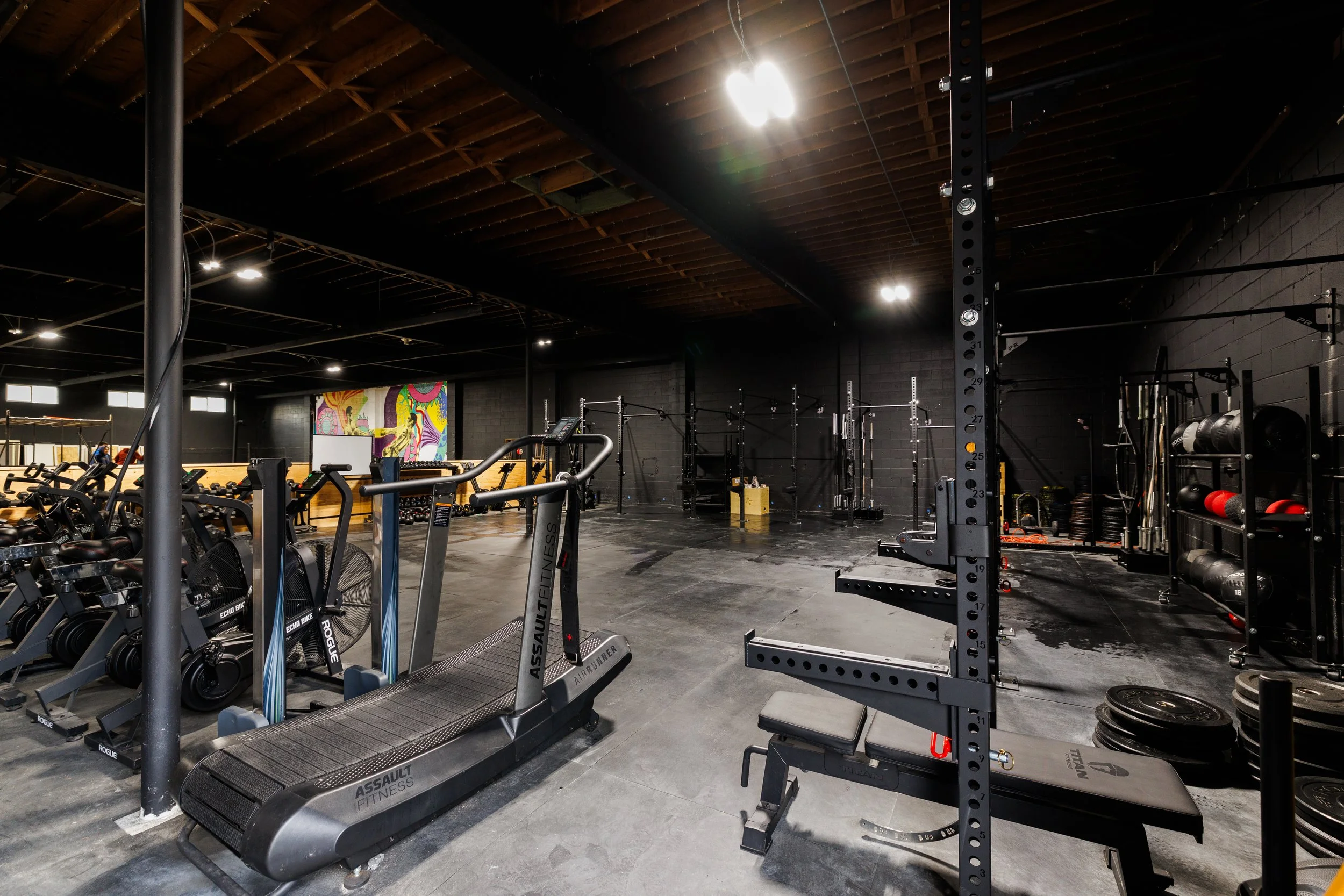 Interior of a modern gym with various workout equipment, including treadmills, bikes, weight racks, and functional training stations, with a colorful abstract mural on the far wall.