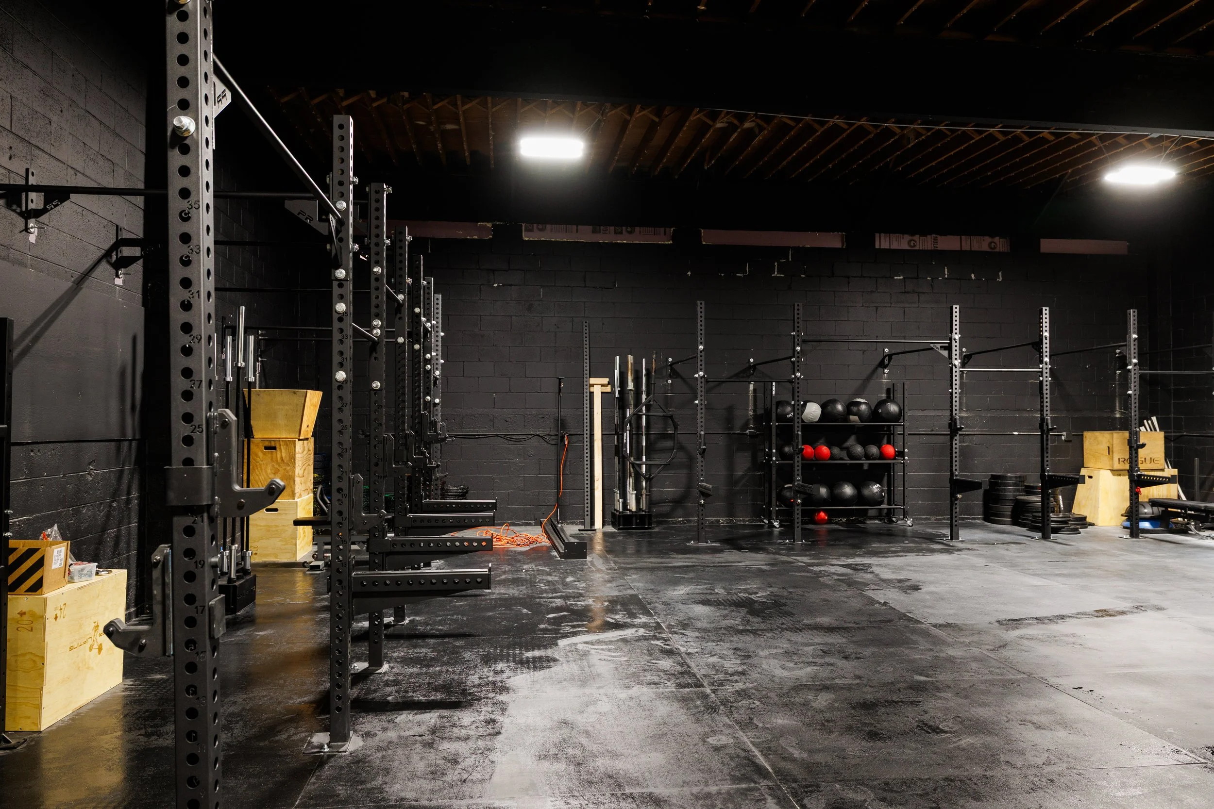 Empty gym with black walls, black flooring, and various weightlifting equipment including squat racks, medicine balls, and plyometric boxes.