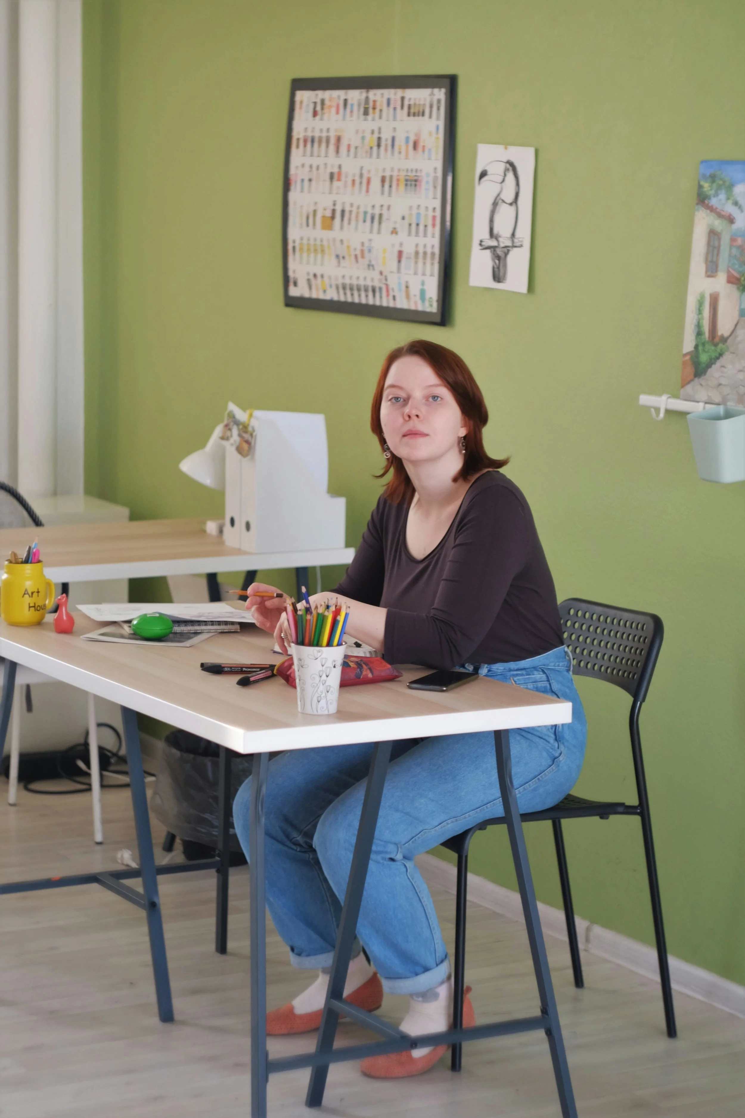 A young woman with red hair and light skin sitting at a white desk in a room with green walls. She is wearing a dark long-sleeve shirt, blue jeans, and orange slippers. The desk has various items including colored pencils in a white cup, a smartphone, and markers. Behind her are framed pictures and paintings on the wall.