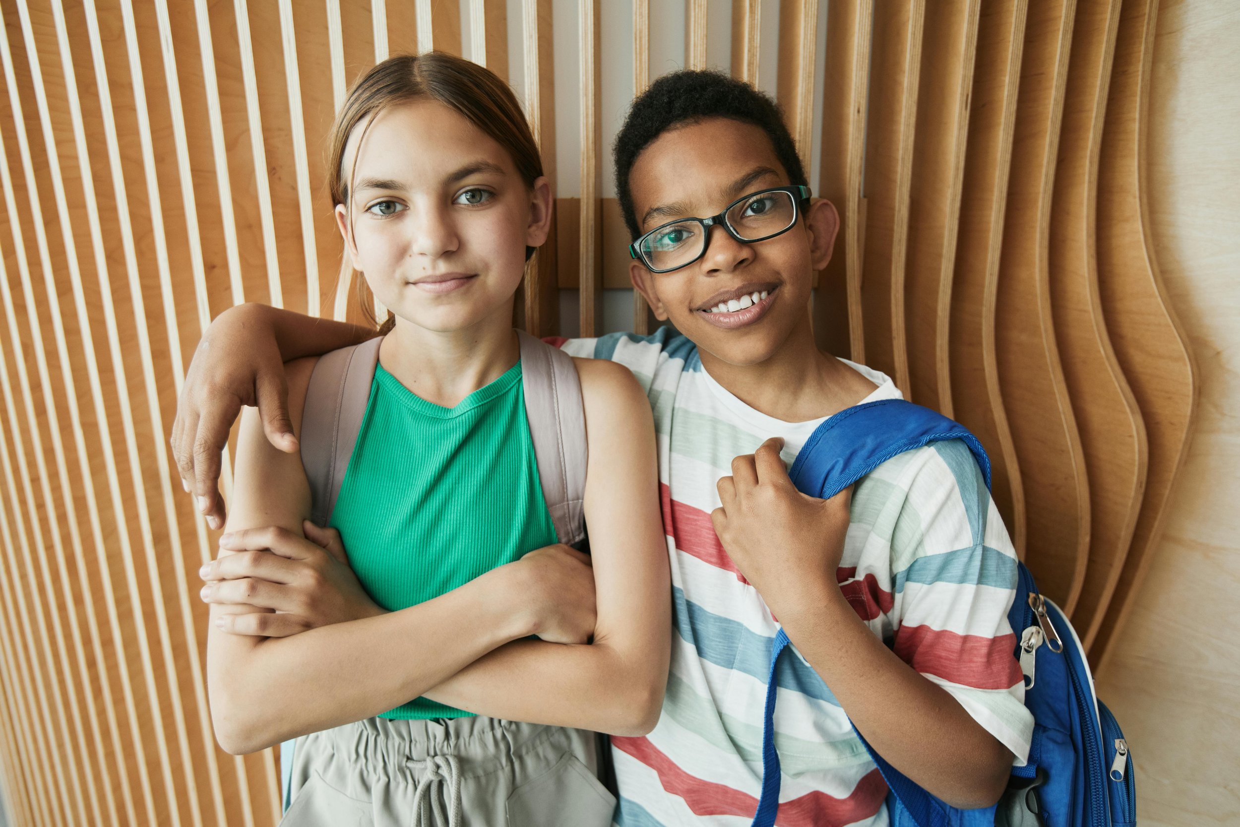 Two children, a girl with blonde hair wearing a green tank top and a boy with glasses wearing a striped shirt, standing close together with arms around each other, smiling in front of a wooden wall.