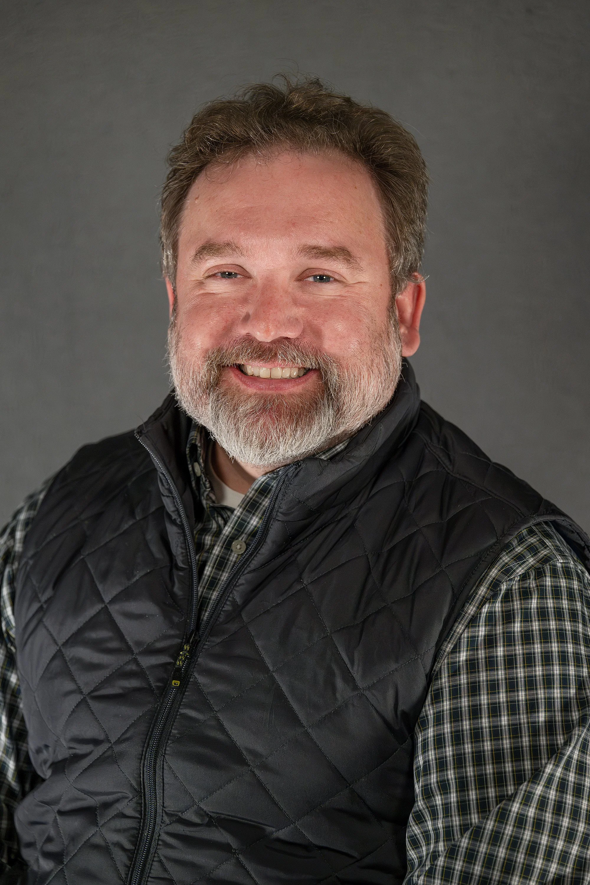 Portrait of a smiling middle-aged man with a beard and mustache, wearing a black quilted vest over a plaid shirt, against a gray background.