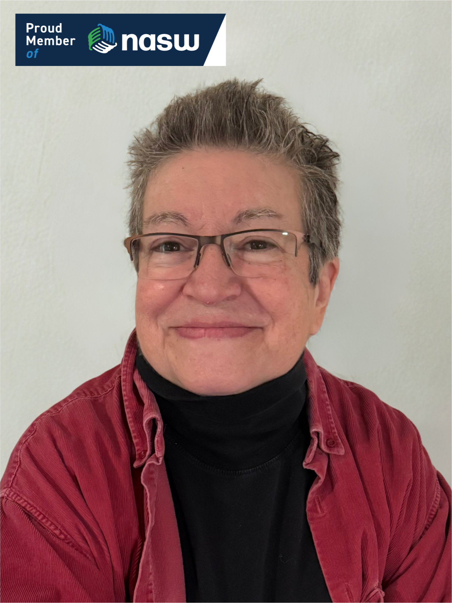 A smiling person with short gray hair, glasses, and a black turtleneck, wearing a red shirt, against a plain light-colored background. There is a NASW badge in the top left corner.