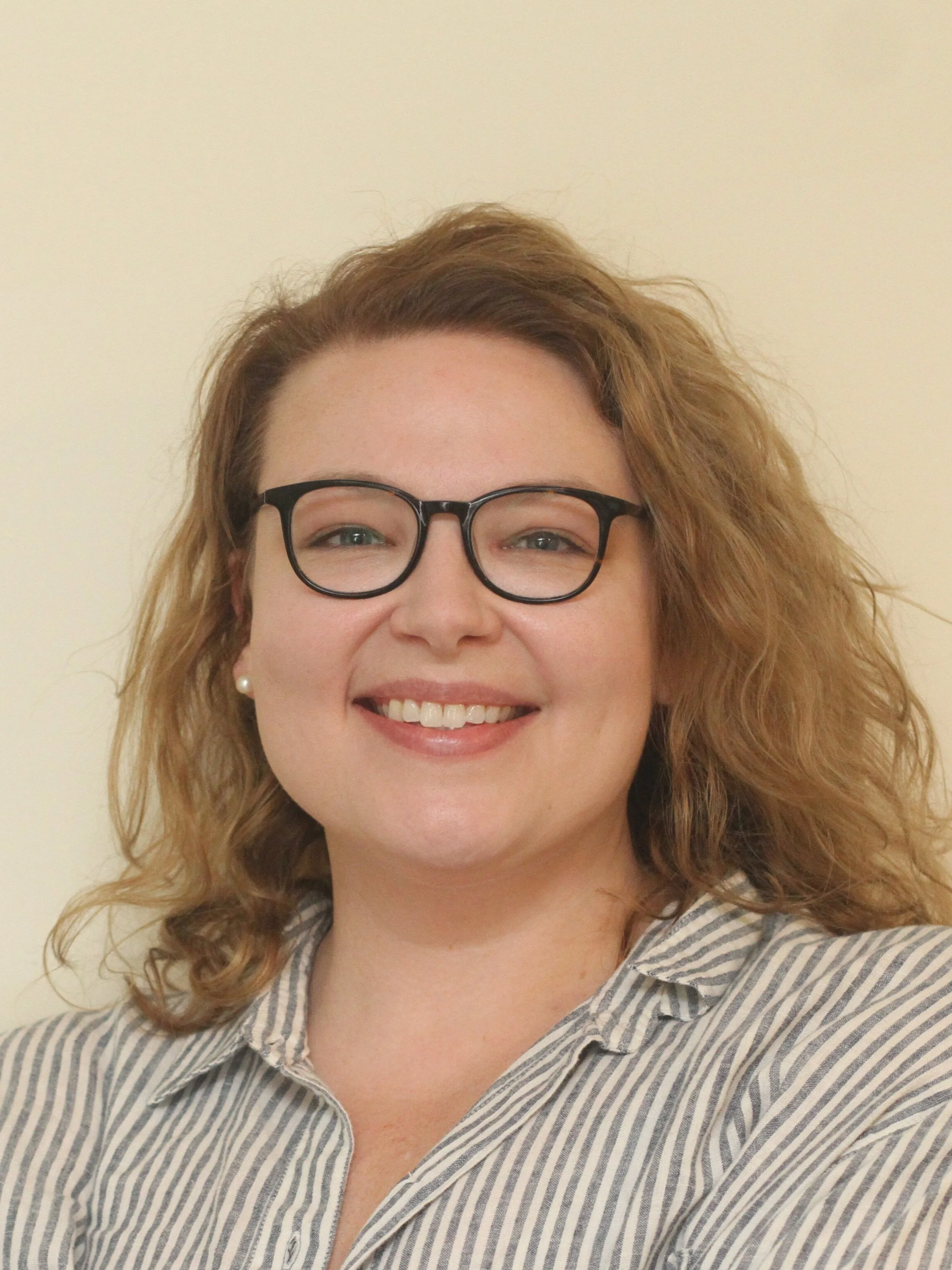 A woman with curly, shoulder-length hair, wearing glasses and a striped shirt, smiling in front of a plain light-colored background.