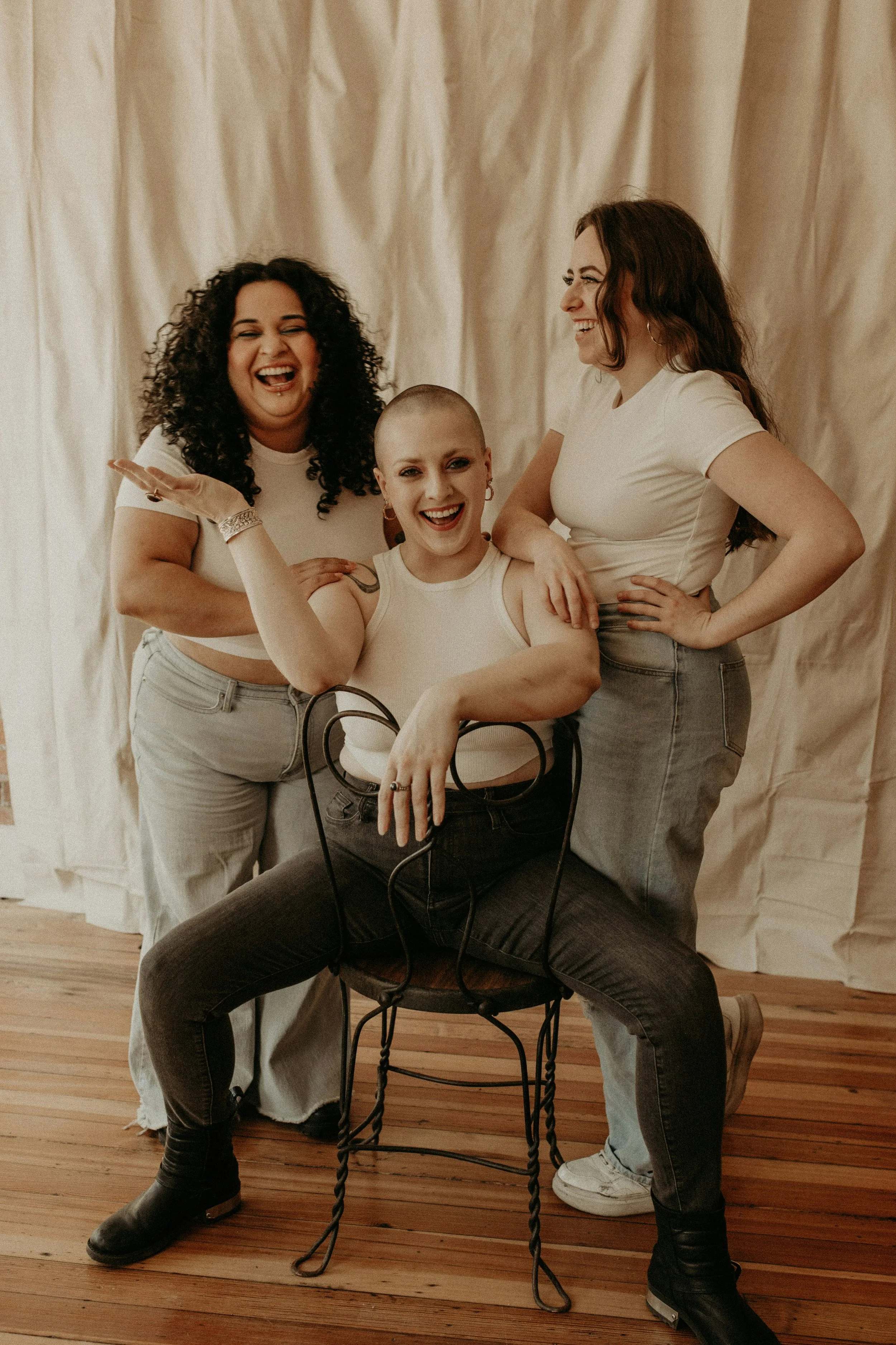 Three women in white tops and gray jeans posing together and smiling, one sitting on a chair with legs spread, one standing and touching her shoulder, and the third standing behind them, all in front of a cream-colored curtain.