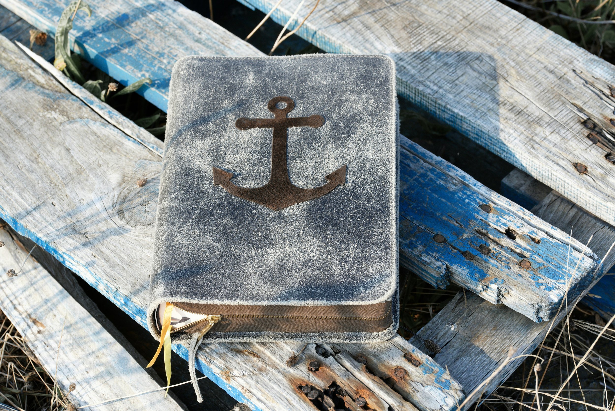 A weathered, sandy-colored notebook with a black anchor symbol on the cover, lying on an old, chipped wooden dock with dried grass nearby.