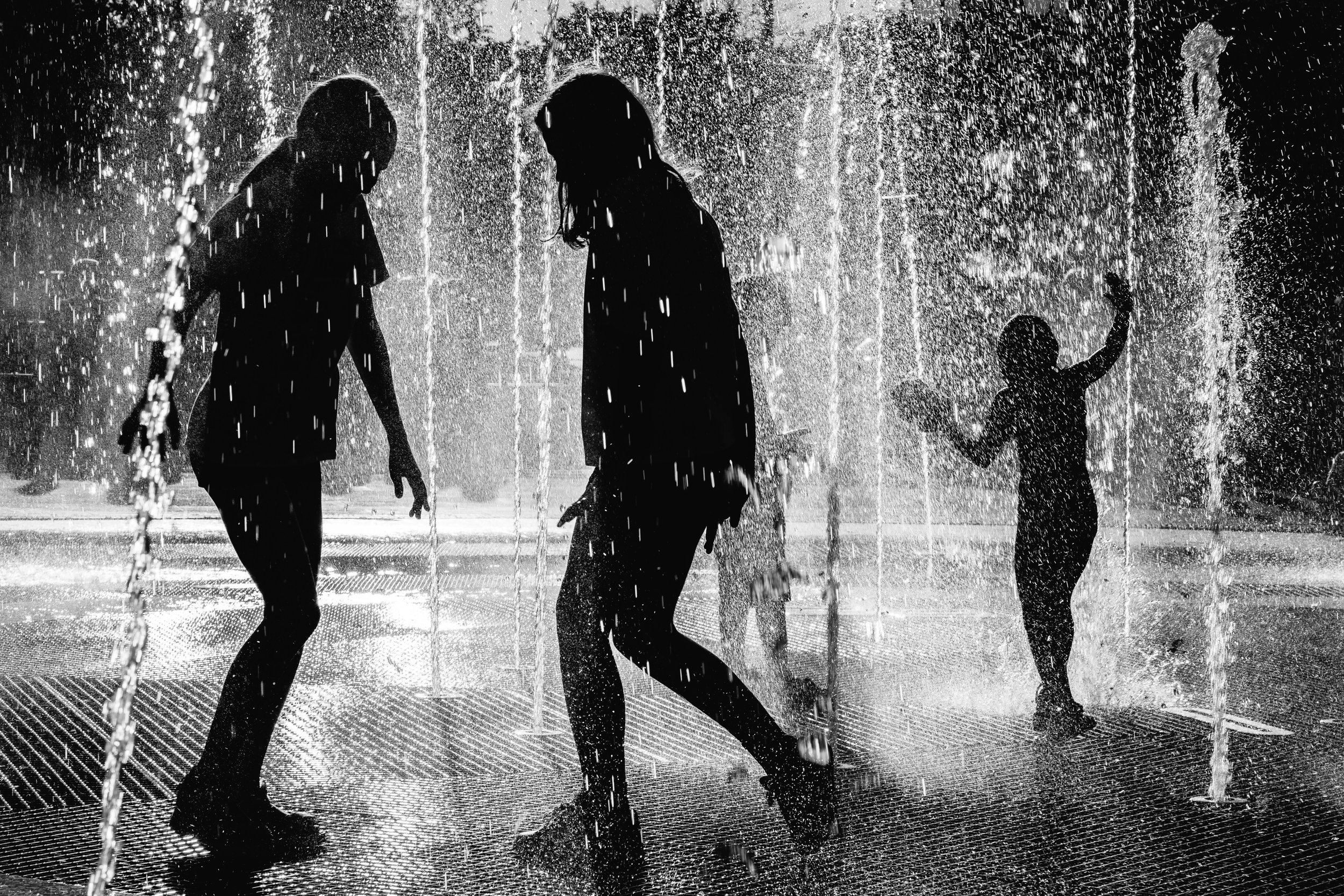 Silhouettes of children playing in a water fountain during rain, with water spraying around them.