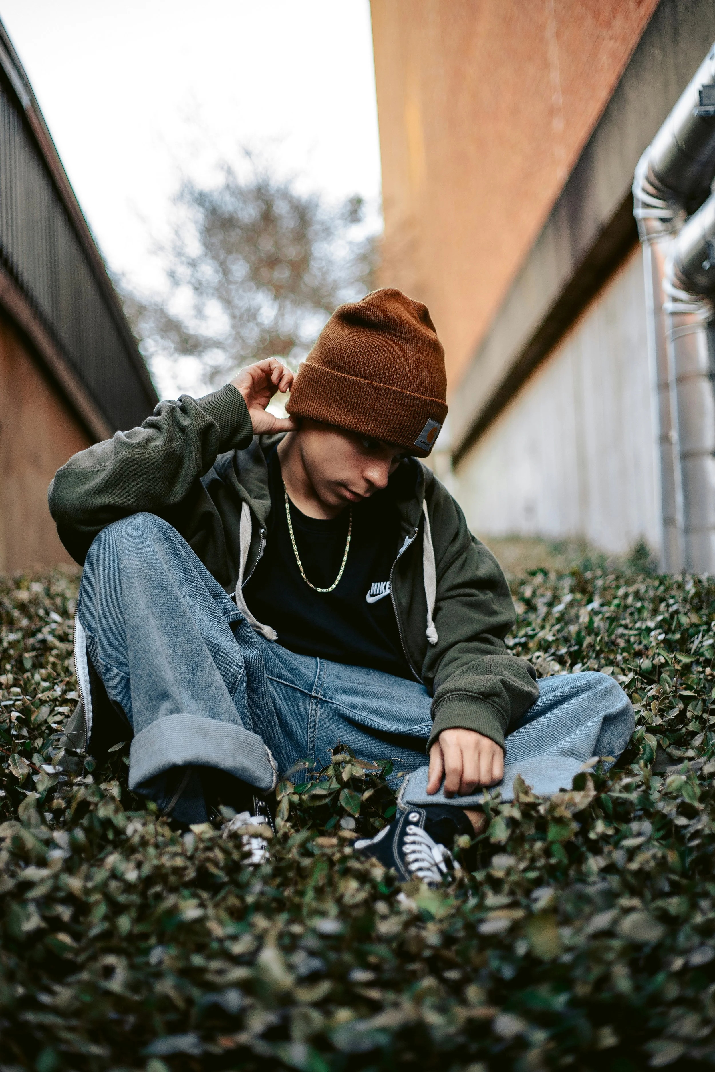 A young man sitting on the ground covered with leaves, wearing a brown beanie, black Nike shirt, olive green jacket, baggy jeans, and black sneakers, adjusting his beanie.