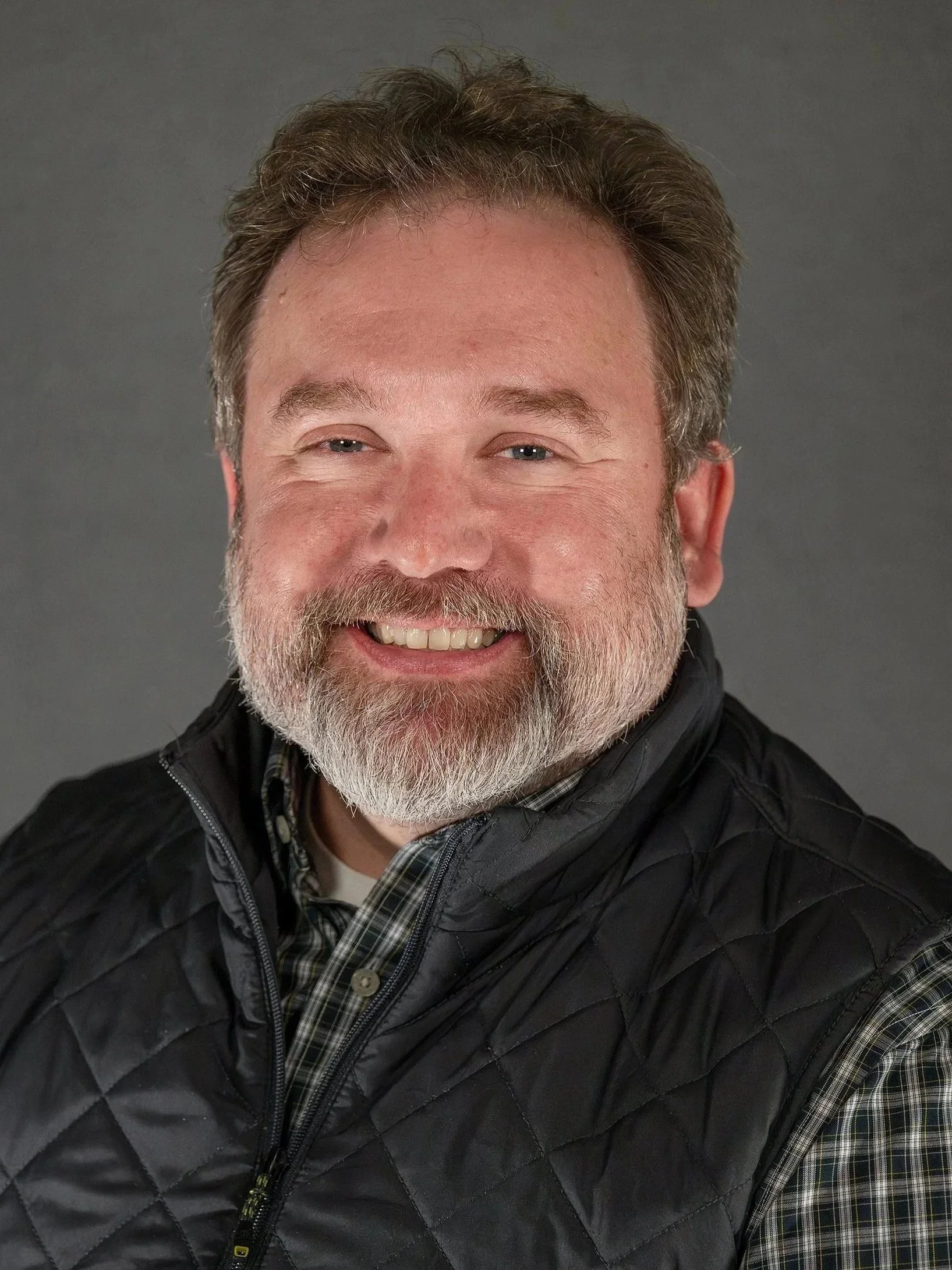 A smiling middle-aged man with a beard and gray hair wearing a black quilted vest over a plaid shirt against a gray background.