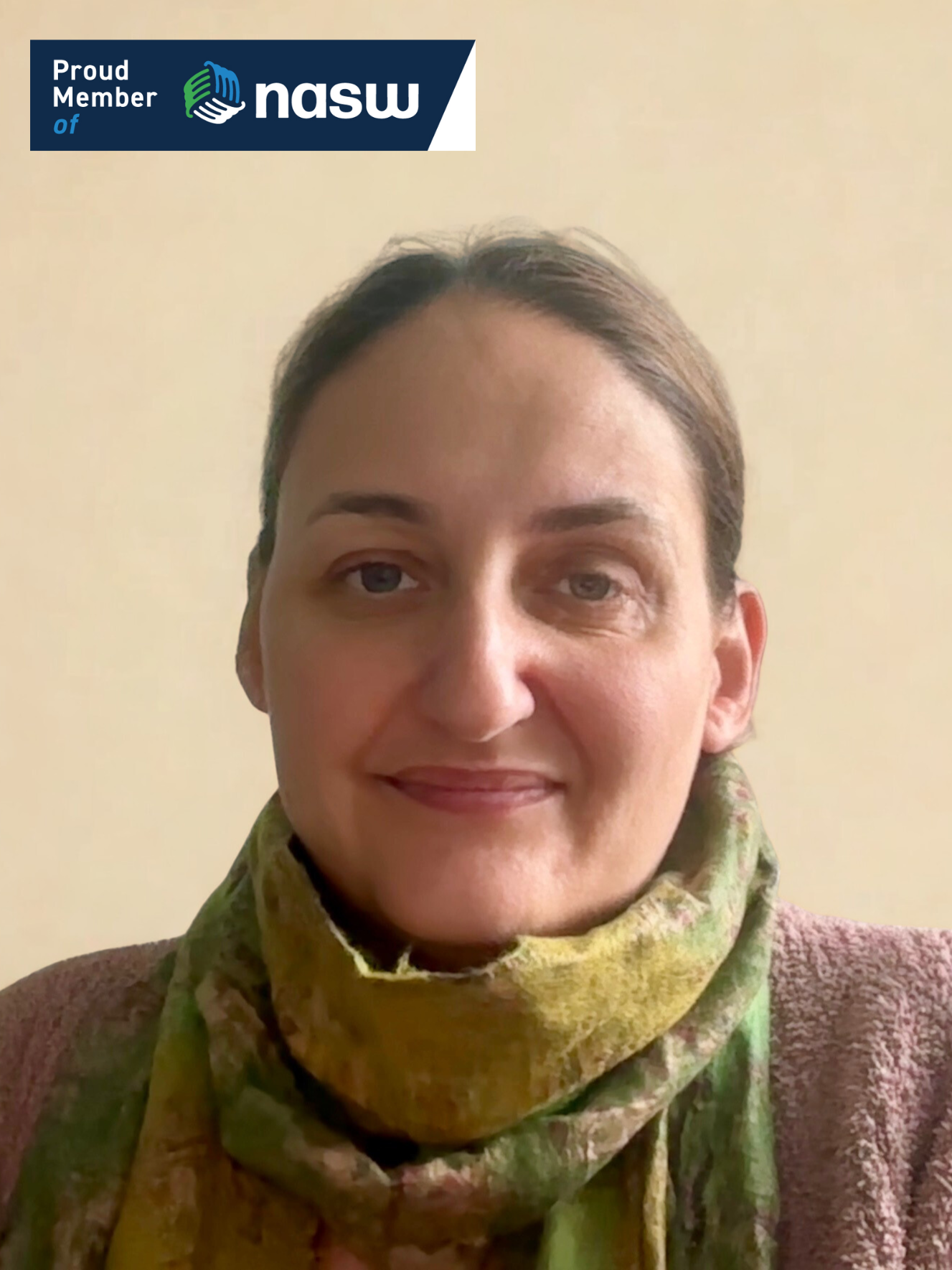 Portrait of a woman with brown hair, wearing a green and yellow scarf and a brown textured blazer, standing against a plain beige background.