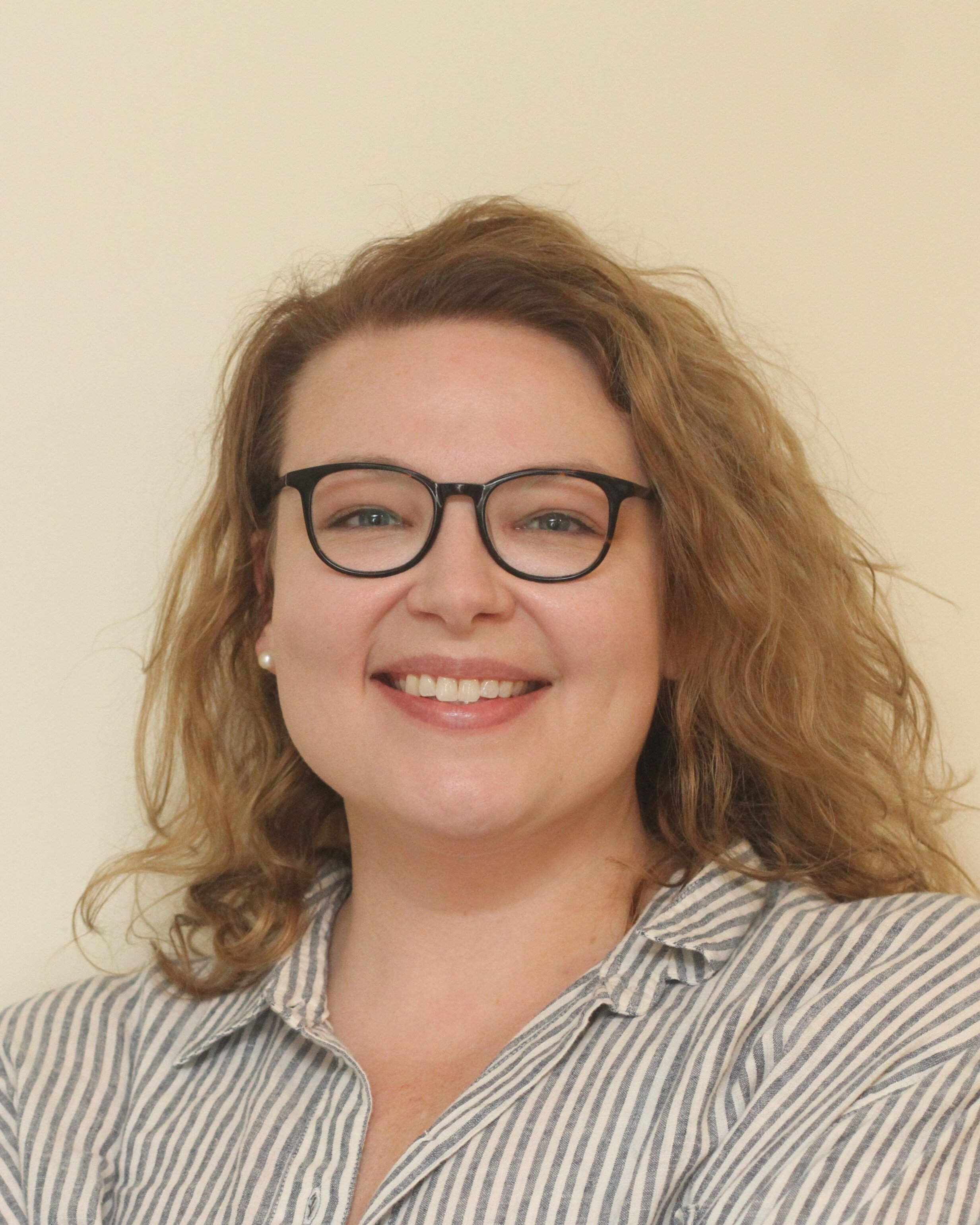 A woman with curly red hair, glasses, pearl earrings, and a striped shirt smiling at the camera.