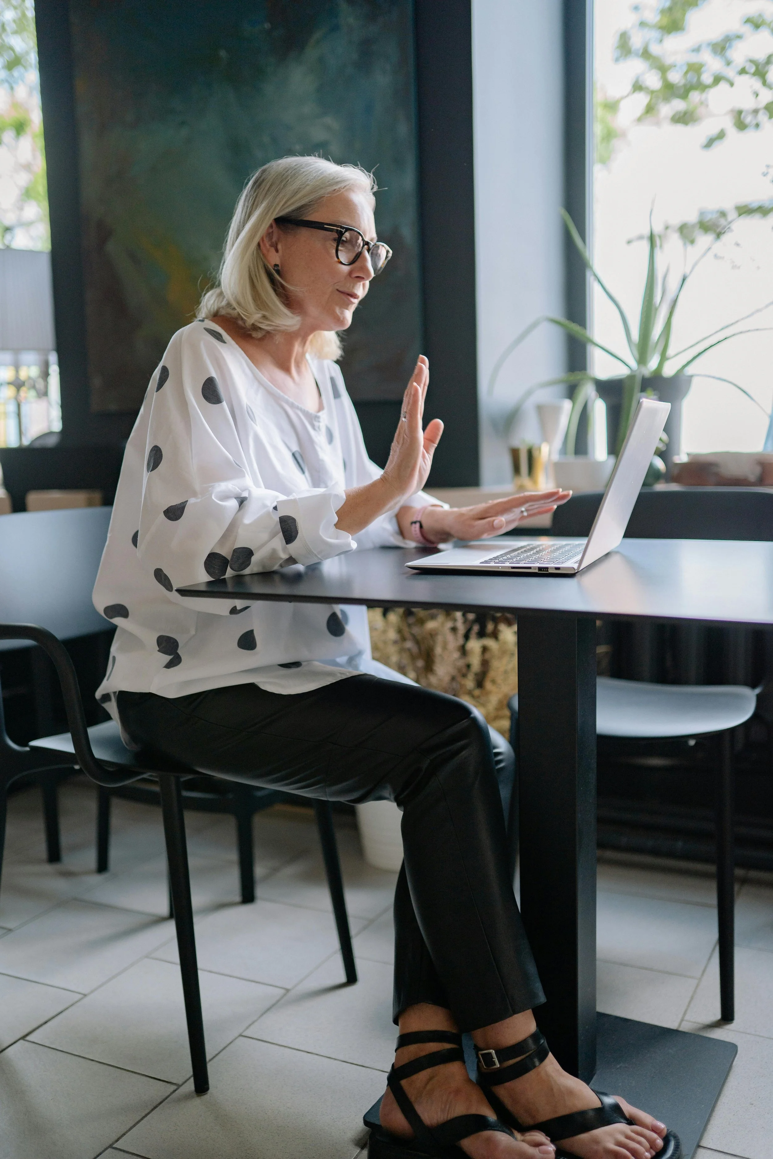 A woman with gray hair, glasses, and a polka-dot shirt having a video call on her laptop in a cafe setting.