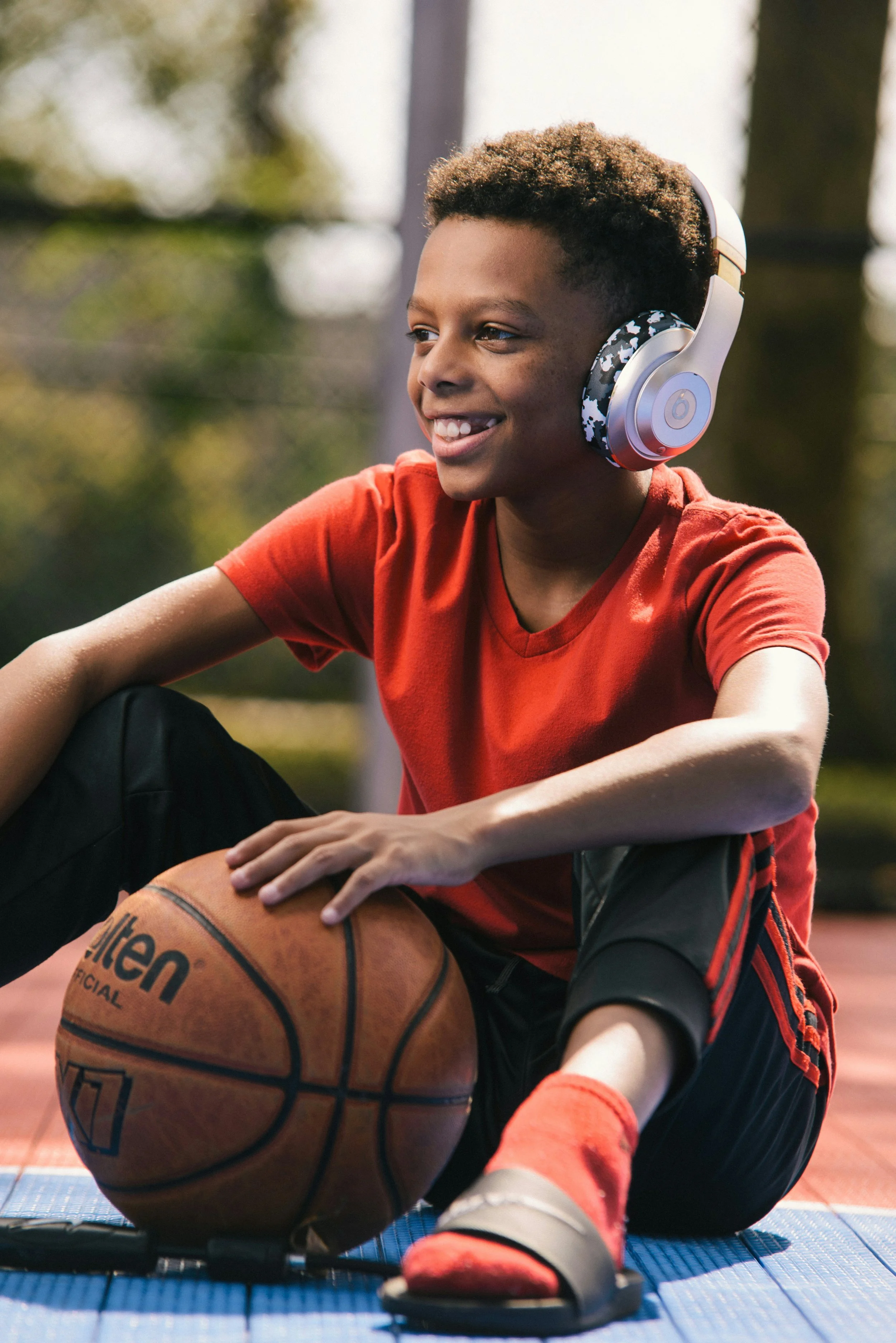 A young boy in a red shirt sitting on a basketball court, holding a basketball, wearing headphones, smiling, with outdoor scenery in the background.