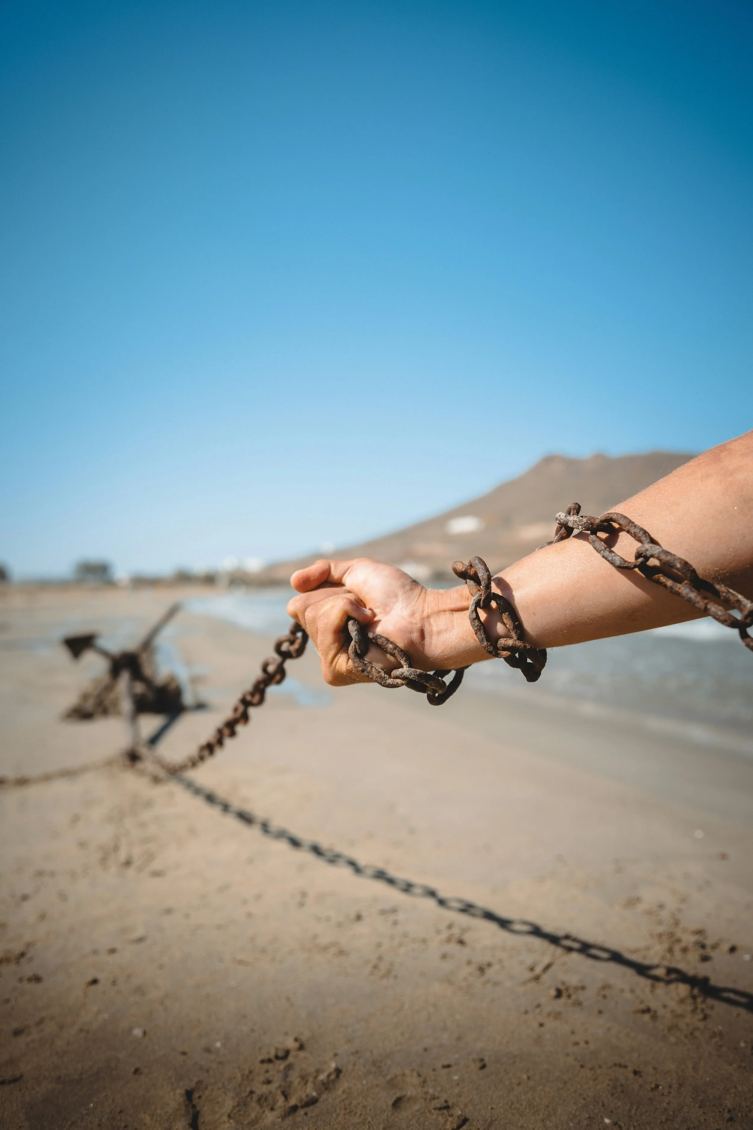A person's arm, wrapped with a chain, pulling an anchor over a sandy beach with the ocean in the background, under a clear blue sky.