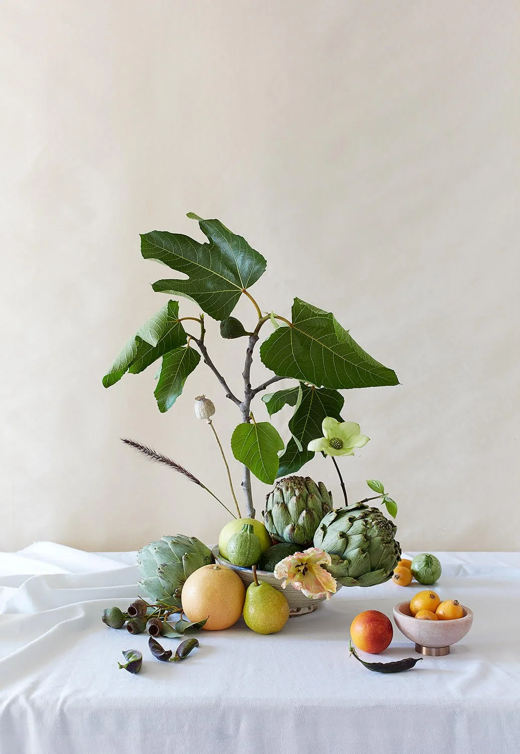 A still life composition of various fruits and greens on a white tablecloth with a plain, light-colored wall in the background.