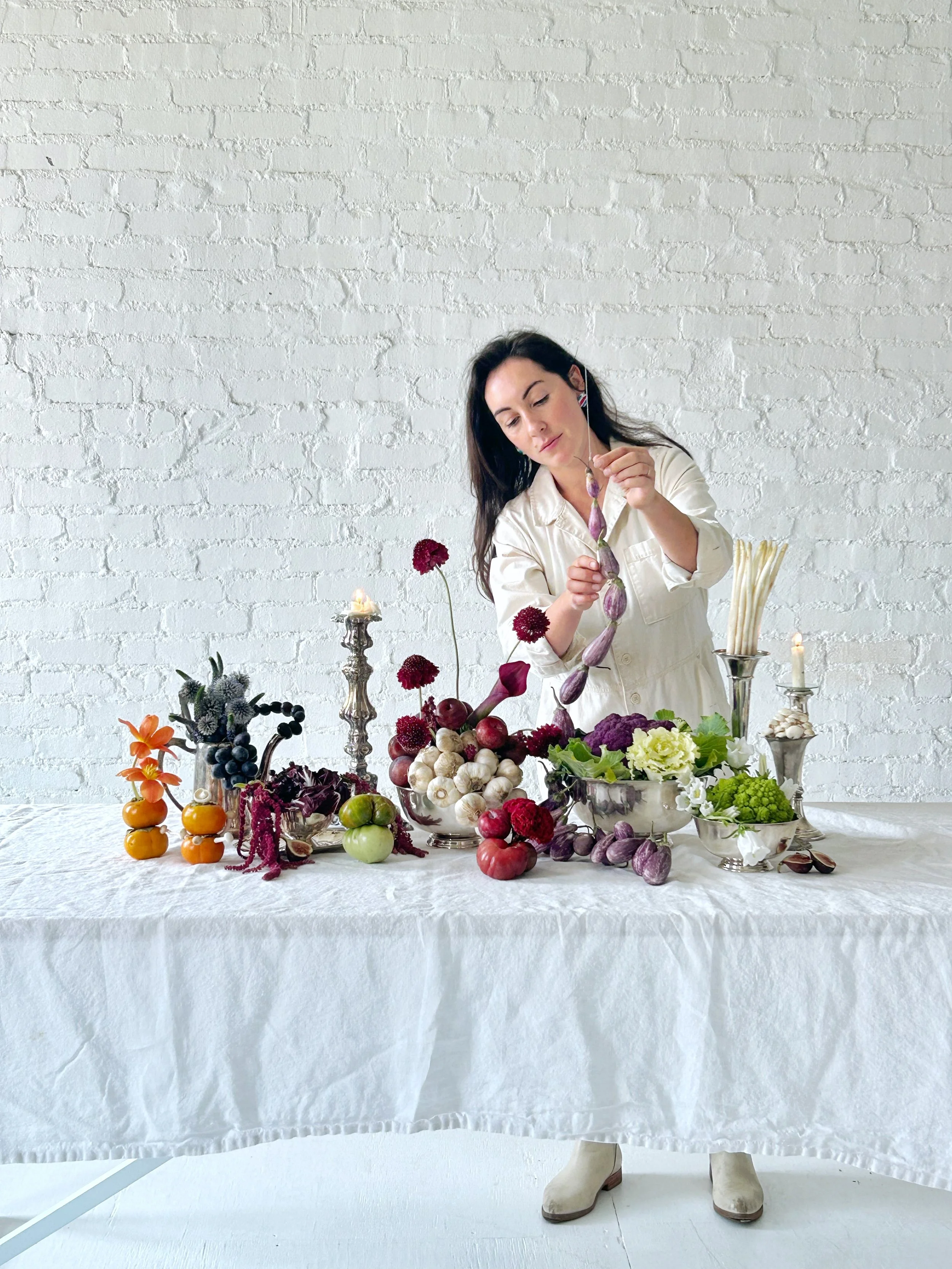 Woman arranging flowers and fruits on a white table against a white brick wall.