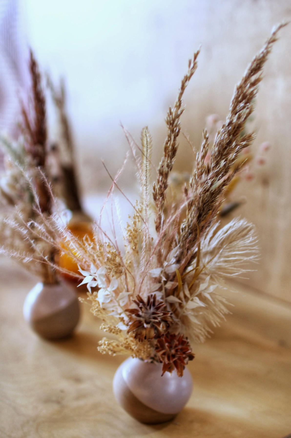 A close-up of a beige ceramic vase containing a dried flower arrangement with various pampas grass, leaves, and seed pods, on a wooden surface.