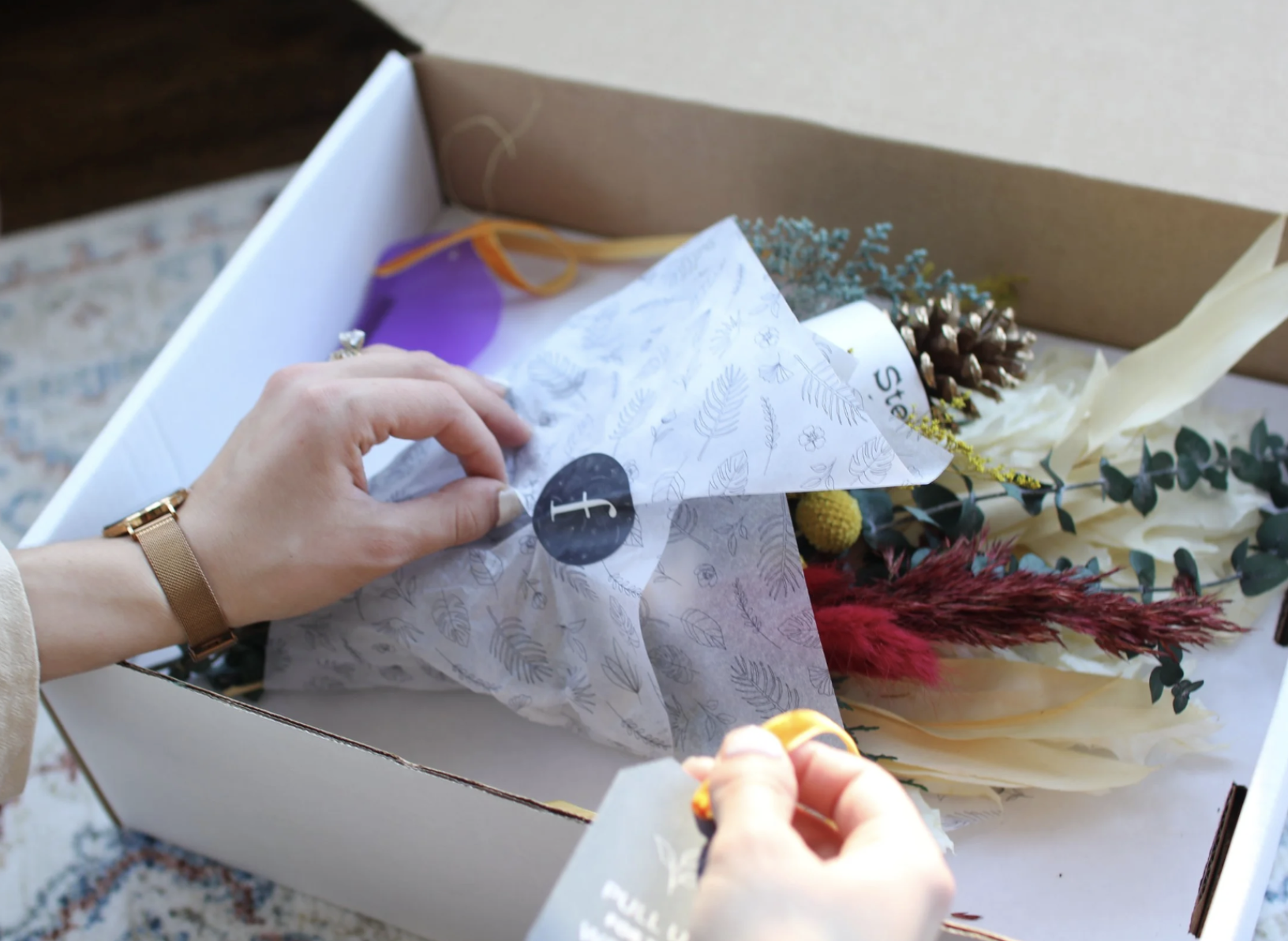 Person wrapping a floral bouquet in tissue paper inside a white box, with various dried and artificial flowers surrounding.
