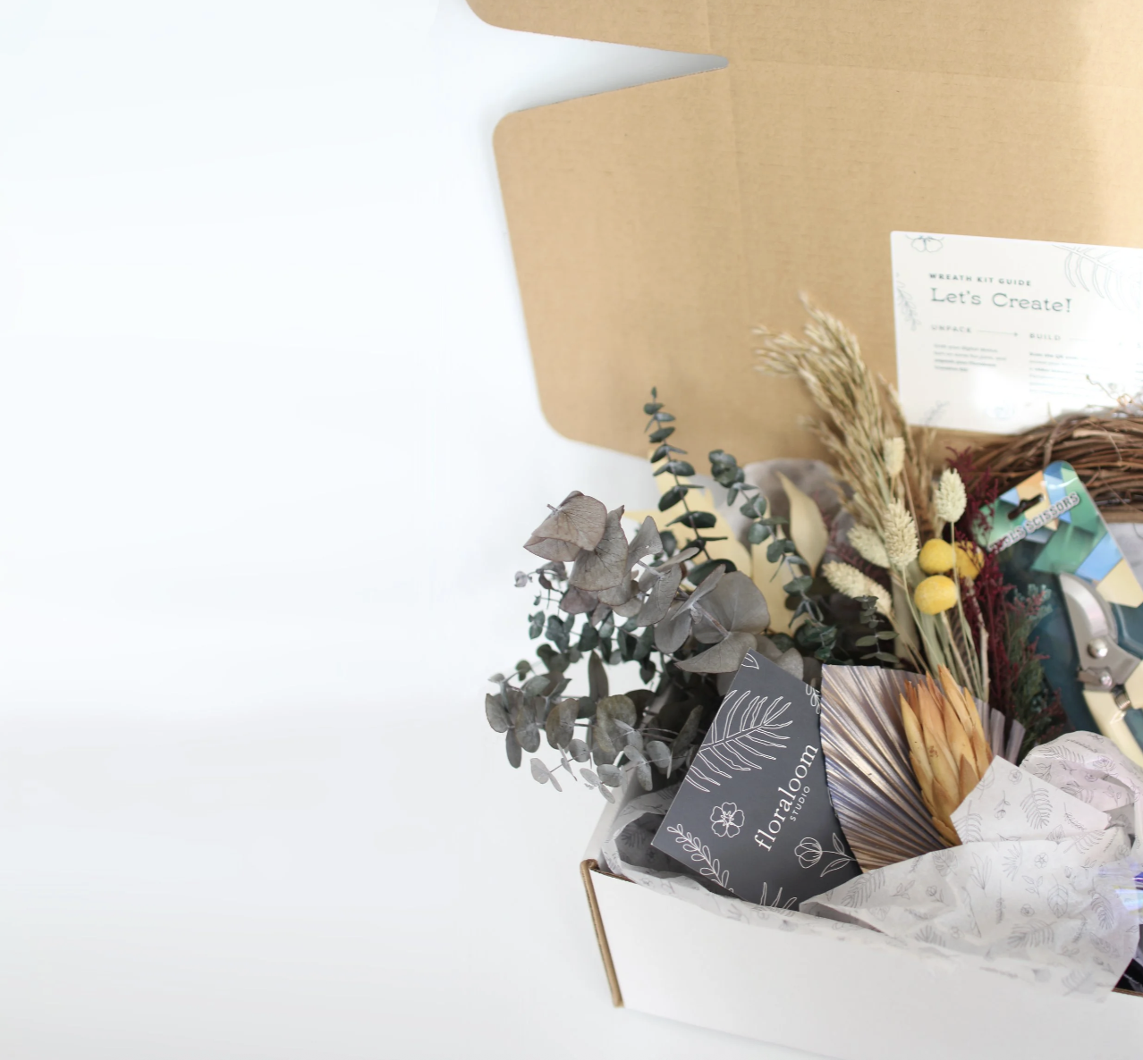 A gift box with dried flowers and foliage, including eucalyptus and wheat, along with floral supplies and an instruction card, all arranged inside a white box with a gold rim.