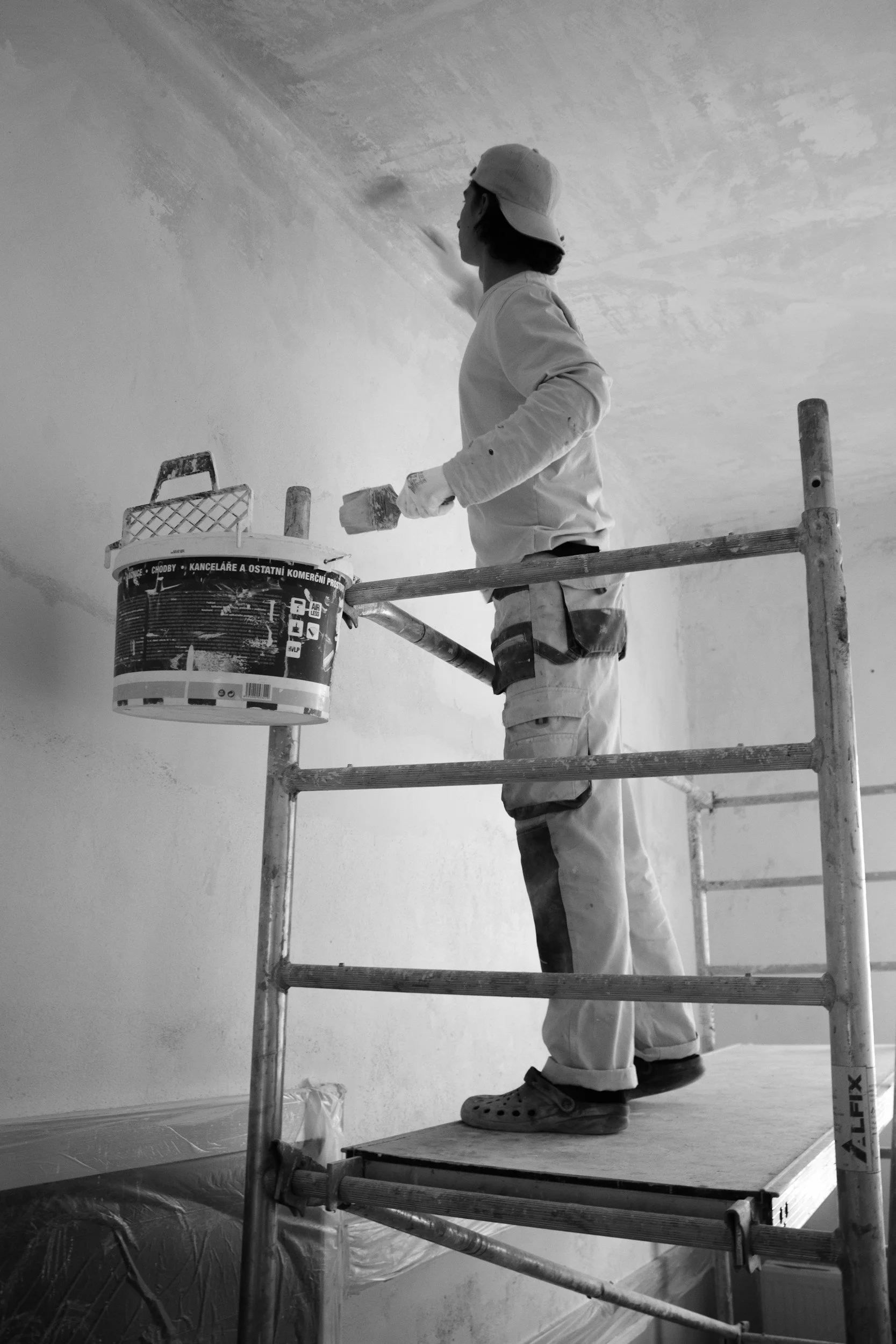 A person wearing a cap and work clothes is standing on scaffolding while painting a wall with a roller brush. There is a bucket of paint attached to the scaffolding.