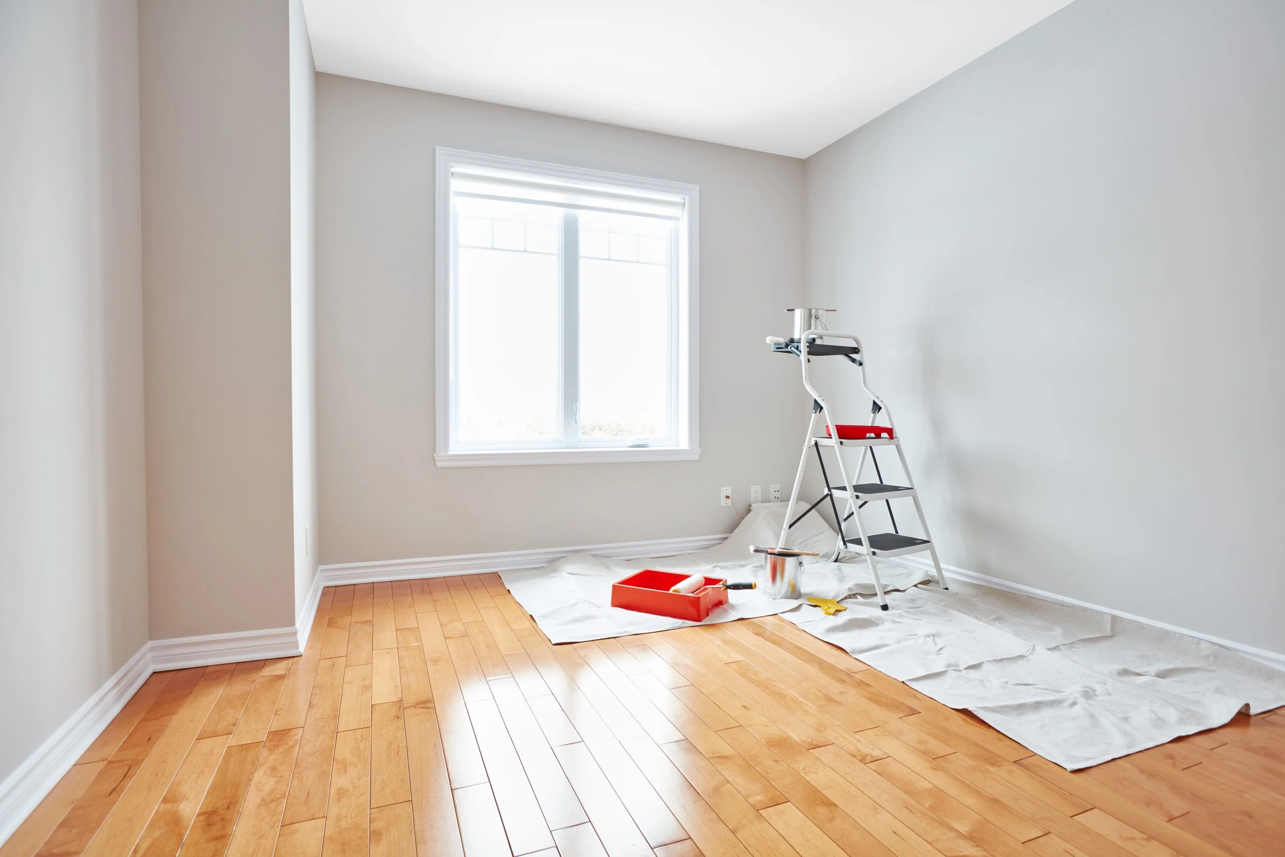Empty room with hardwood floors being painted or renovated, paint supplies on the floor, a napkin under the supplies, a window with natural light, white walls, a ladder with some tools on it.