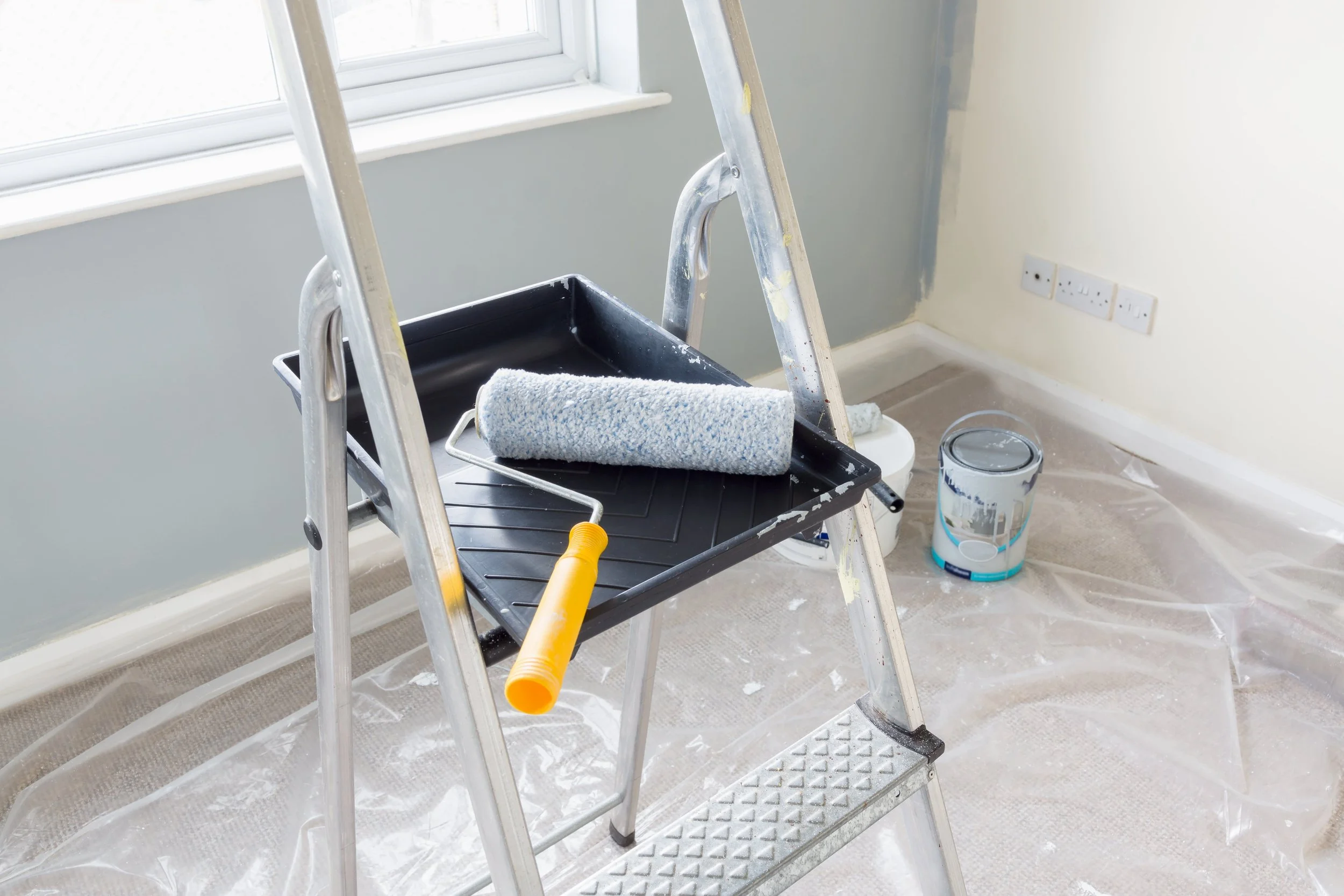 Room under renovation with ladder, paint roller, paint tray, and a bucket of paint, plastic sheeting on the floor, and electrical outlets on the wall.