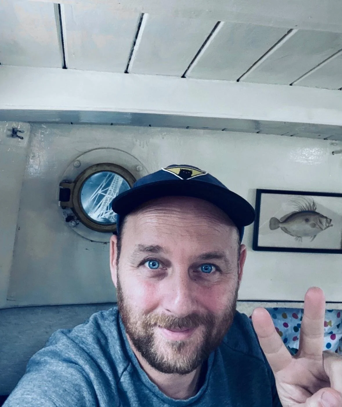 A man with blue eyes and a beard taking a selfie, wearing a dark cap with a yellow emblem. In the background, there is a fish sketch on the wall and a porthole window reflecting a bridge.