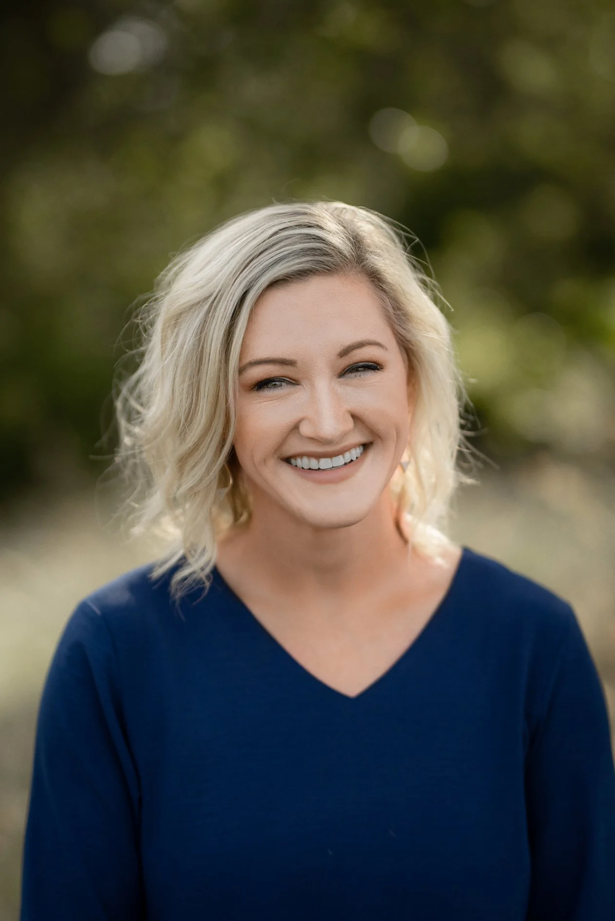 A smiling woman with platinum blonde, shoulder-length curly hair, wearing a navy blue V-neck top, outdoors with blurred green foliage in the background.