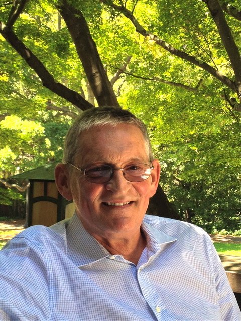 Smiling older man with glasses in a white shirt outdoors with green trees and sunlight in the background.