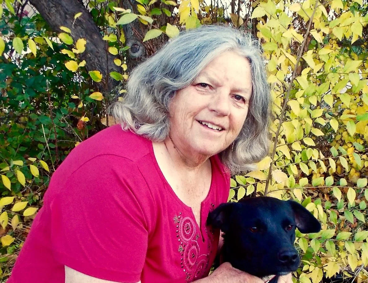 An elderly woman with gray hair wearing a pink shirt, smiling and holding a black dog outdoors. There are yellow and green leaves on bushes behind her.