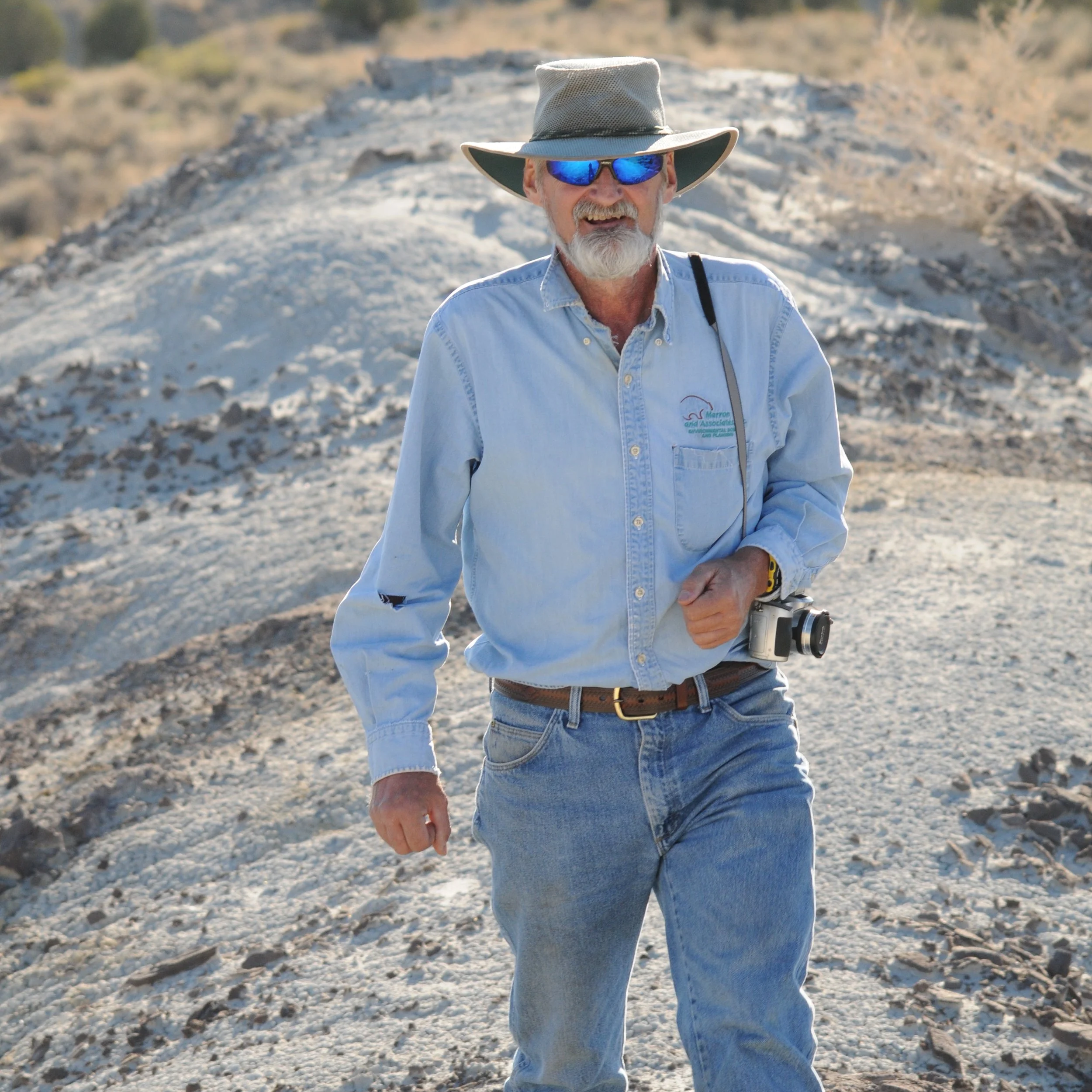 An older man with a beard wearing a wide-brimmed hat, sunglasses, a long-sleeved blue shirt, and jeans, standing outdoors in a rocky, desert-like area with hills in the background.