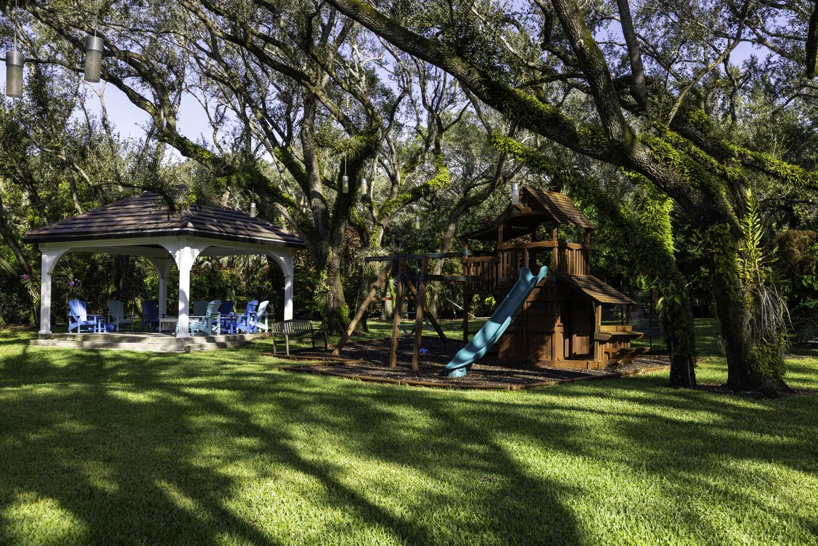 A lush backyard with a wooden playhouse, slide, swings, and a shaded seating area under large trees with hanging lanterns.
