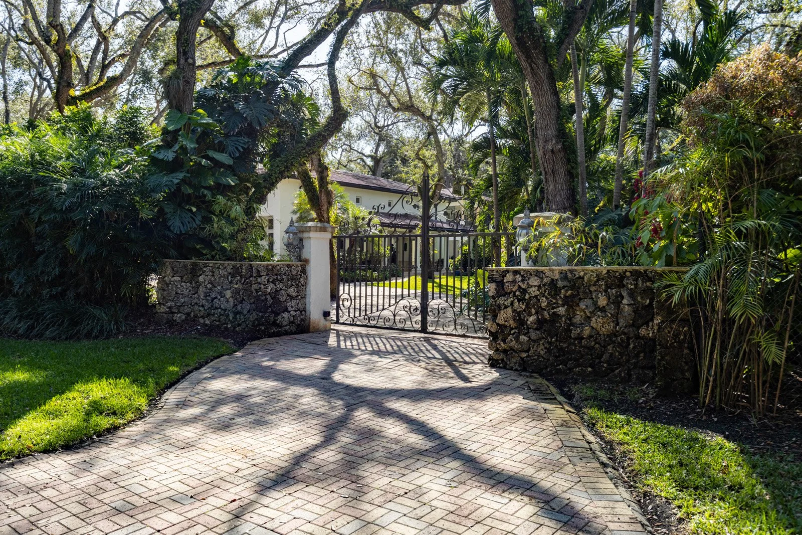 Wrought iron gate and stone wall entrance to a lush, landscaped property with brick driveway, tall trees, and tropical plants.