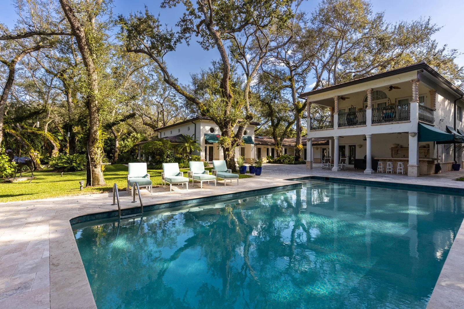 A backyard swimming pool surrounded by lounge chairs, trees, and a two-story house with a patio and balcony.