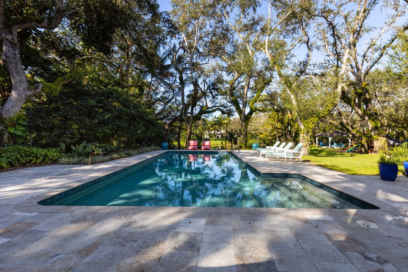 A backyard with a swimming pool surrounded by trees and outdoor seating including white lounge chairs and colorful chairs, with a grassy area and a gazebo in the background.