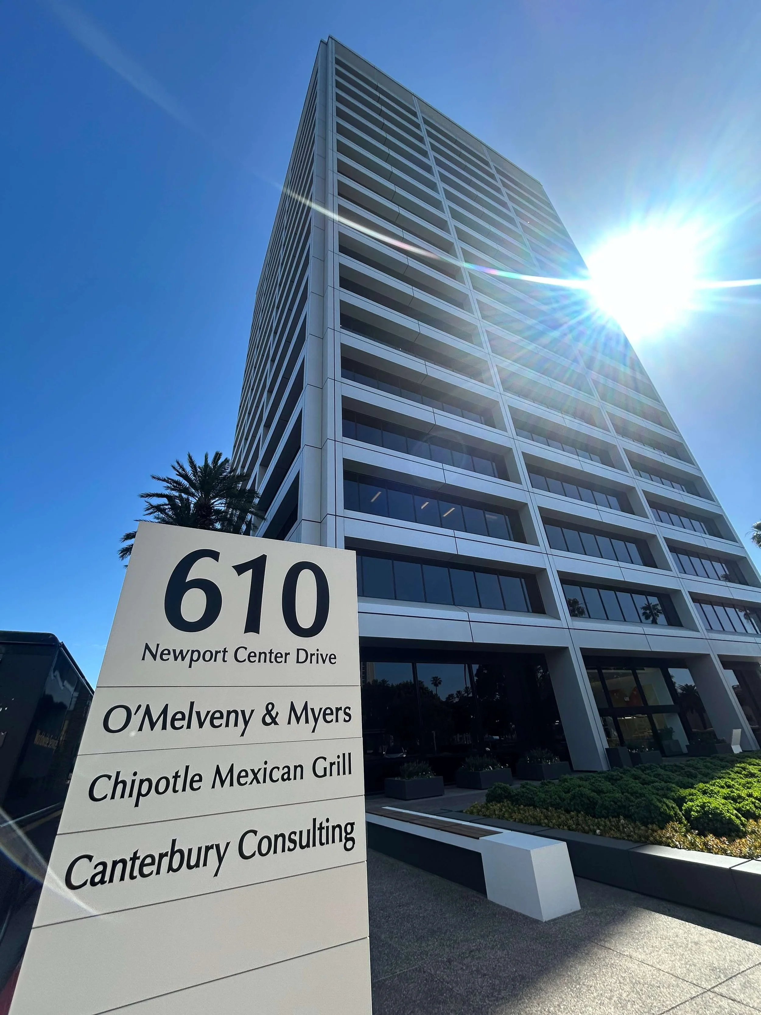 610 Newport Center Drive Suite 250, Newport Beach, CA 92660.  The office building location for Tenant Eviction Defense.  Tall modern office building with a blue sky and bright sun, and a white sign with black text listing the address and tenants.