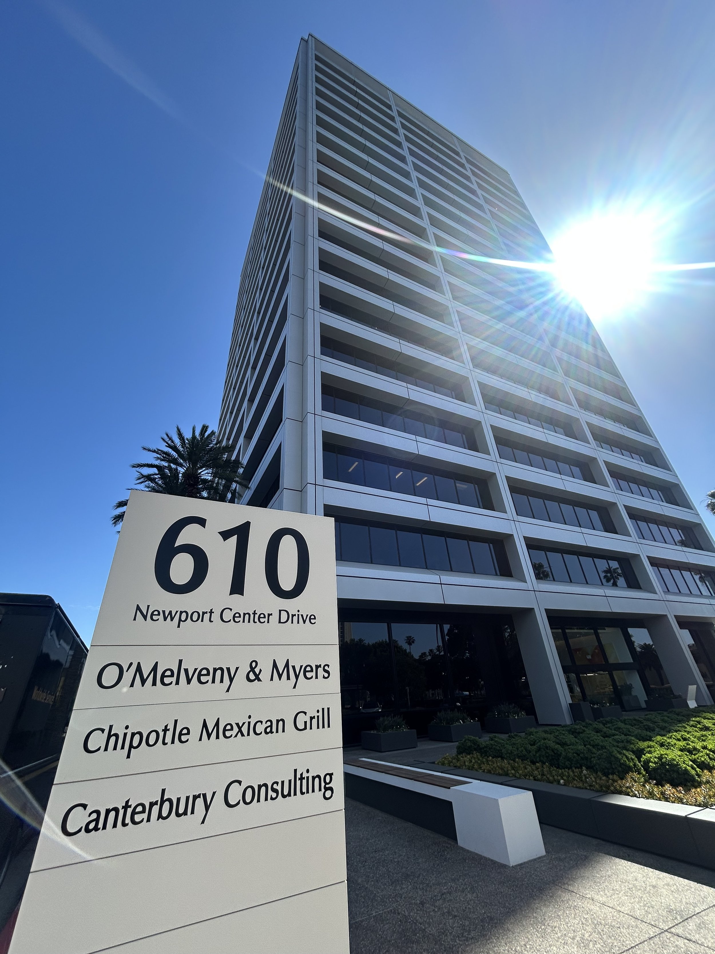 A tall modern office building with a reflective glass facade under a bright blue sky, with the sun shining brightly. A white sign in the foreground displays the address 610 Newport Center Drive and lists tenants including O'Melveny & Myers, Chipotle Mexican Grill, and Canterbury Consulting.