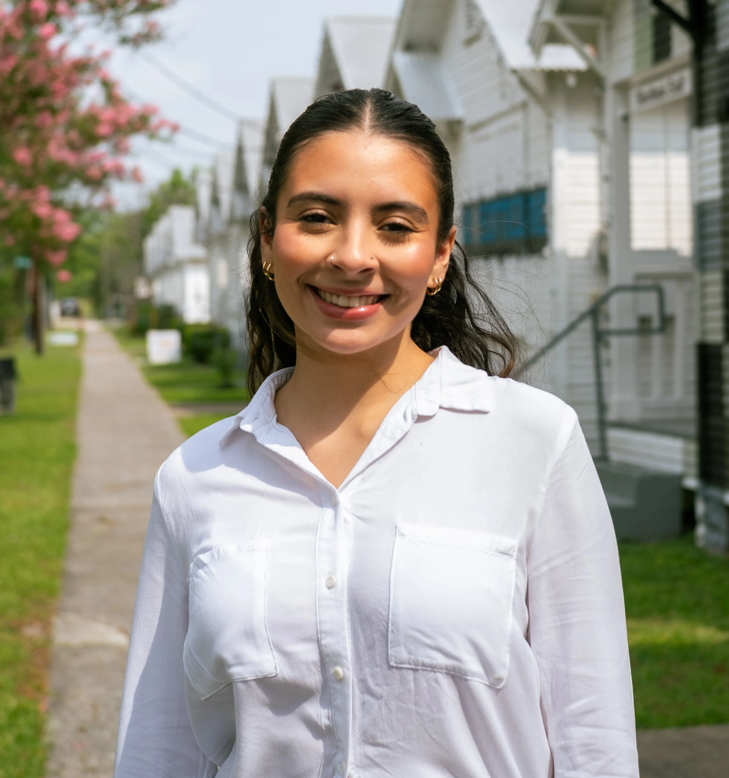 A smiling woman with dark wavy hair pulled back, wearing a white button-up shirt, standing on a sidewalk in a residential neighborhood with white houses, green lawns, and trees with pink blossoms.