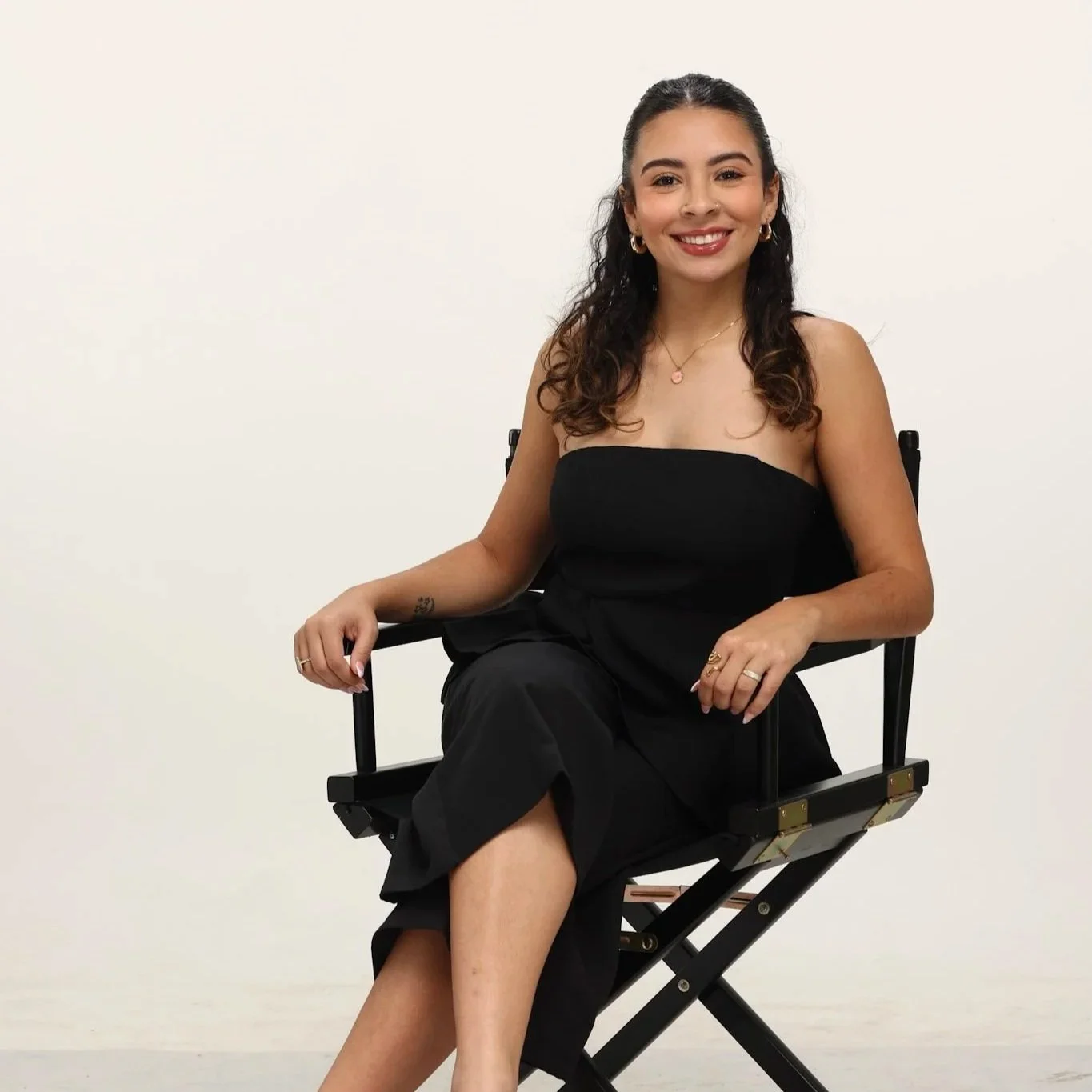 A woman with curly dark hair, wearing a strapless black top and black pants, sitting on a black director's chair against a plain white wall, smiling at the camera.