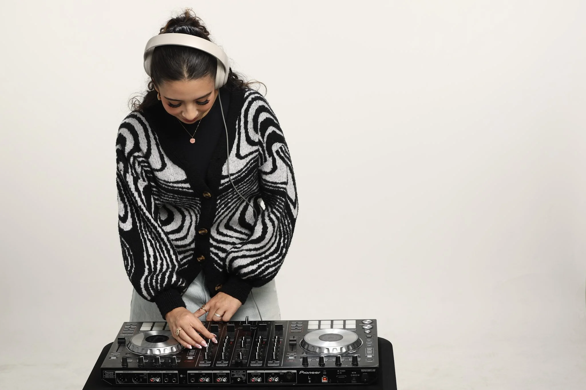 A woman wearing a black and white patterned sweater and large headphones is DJing on a Pioneer mixer, with a plain white wall in the background.