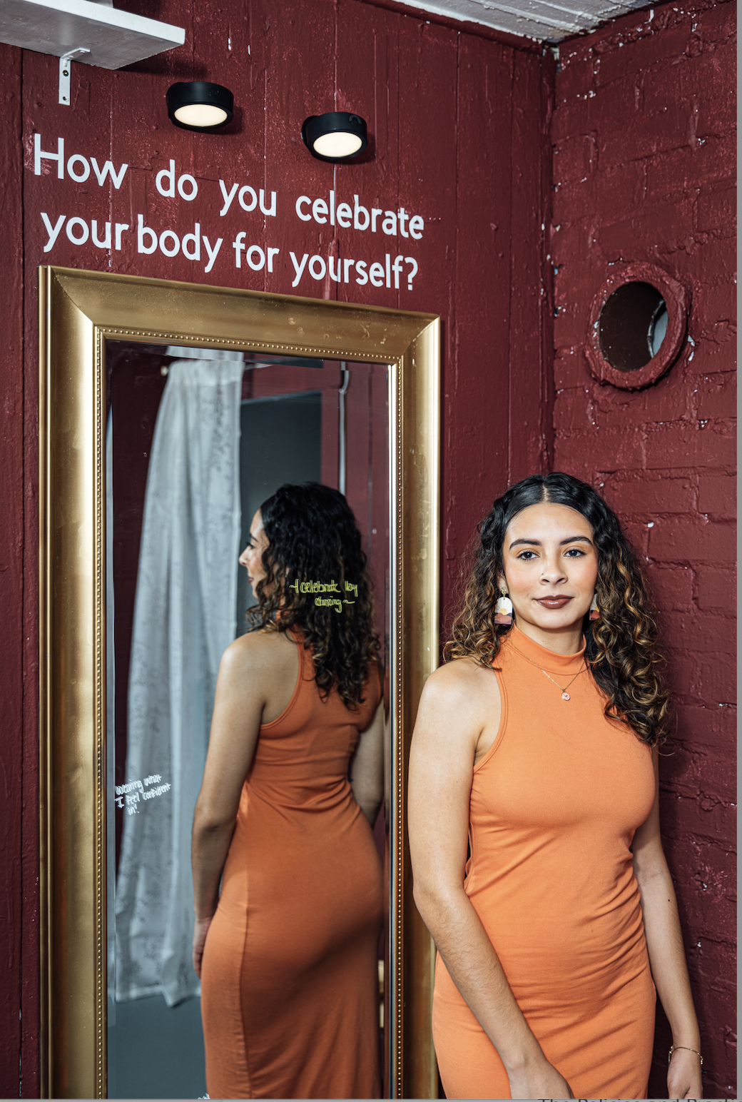 A woman with curly hair in an orange dress standing next to a mirror in a red room. The mirror shows her back and a reflection of her side. There is a sign on the wall that reads, "How do you celebrate your body for yourself?"
