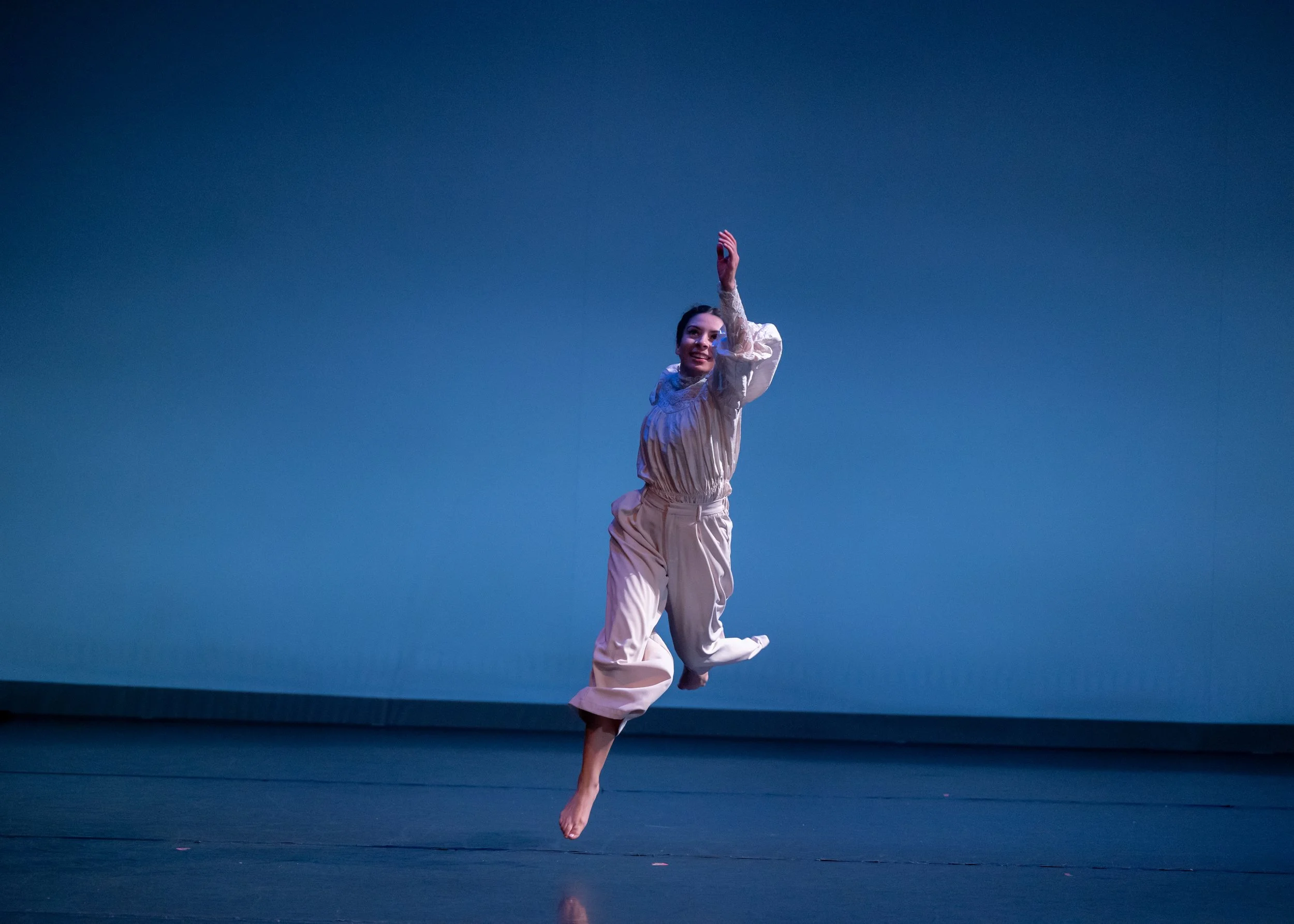 A female dancer in a cream-colored outfit mid-air on a stage with a blue background.