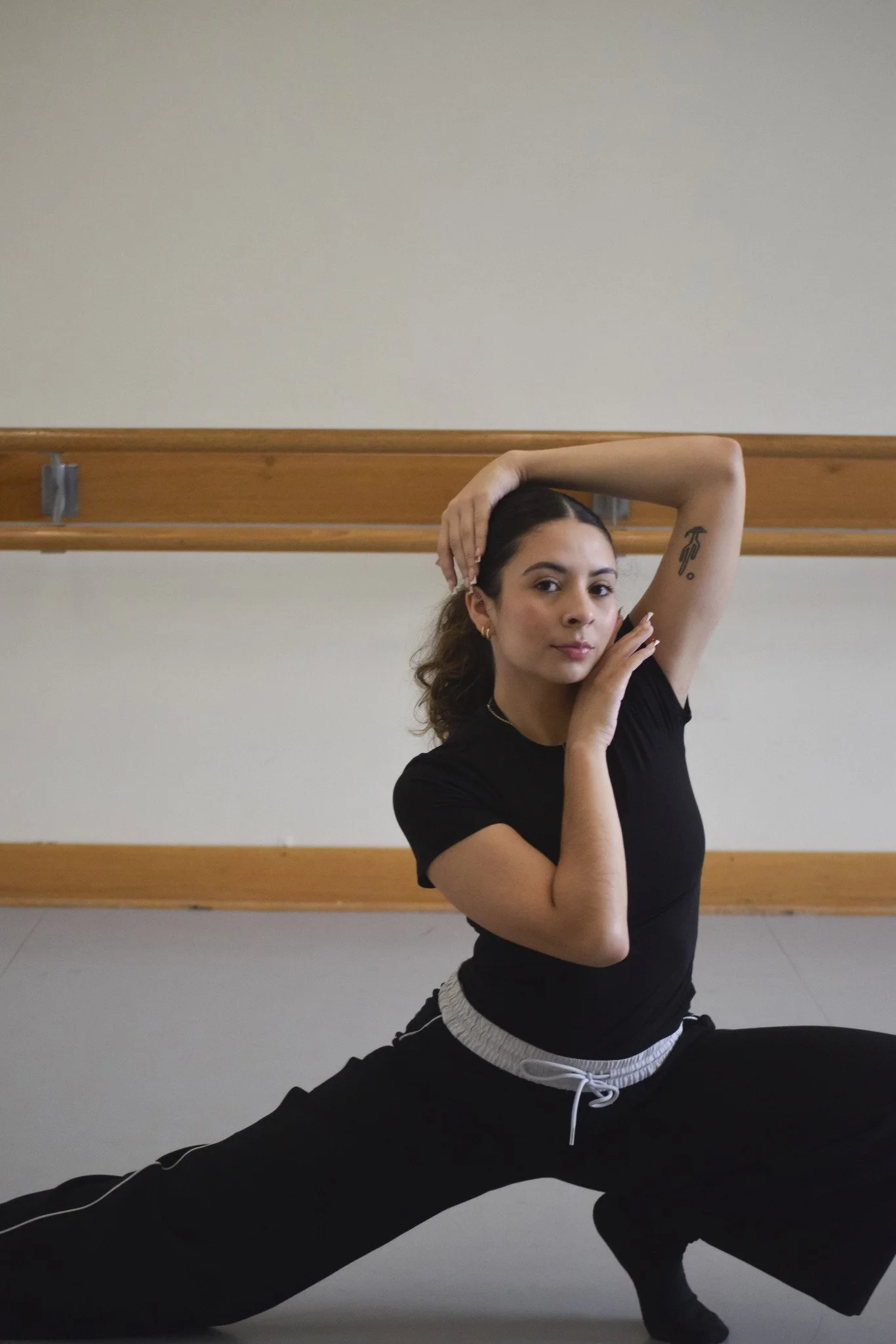 Woman in black athletic wear stretching on the floor in front of a ballet barre, with one arm raised and resting on her head, performing a split pose.