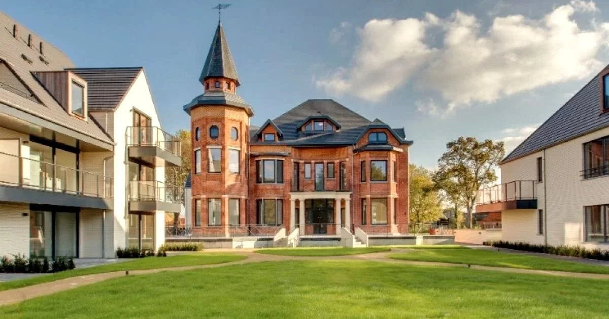 Een groot, indrukwekkend huis met een toren, gebouwd van rood baksteen, omgeven door moderne witte appartementen met balkons en een groene tuin. De lucht is blauw met enkele wolken.