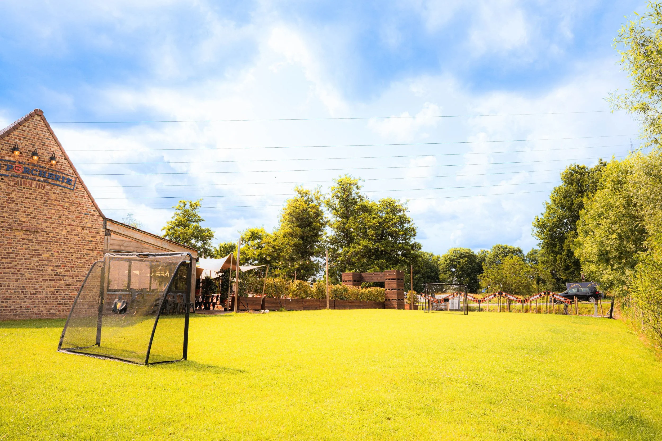 Een grasveld met een klein voetbaldoeltje, omgeven door bomen en een gebouw met een bord 'De Porcherie' op de gevel, onder een blauwe hemel met wat wolken.