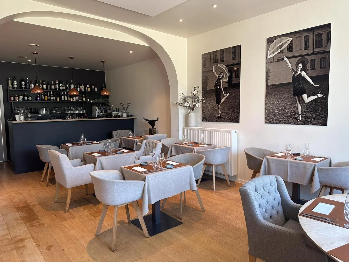 Interieur van een restaurant met houten vloeren, beige stoelen en tafels met wijn- en waterglazen. Aan de muur hangen twee zwart-wit foto's van vrouwen met parasols.