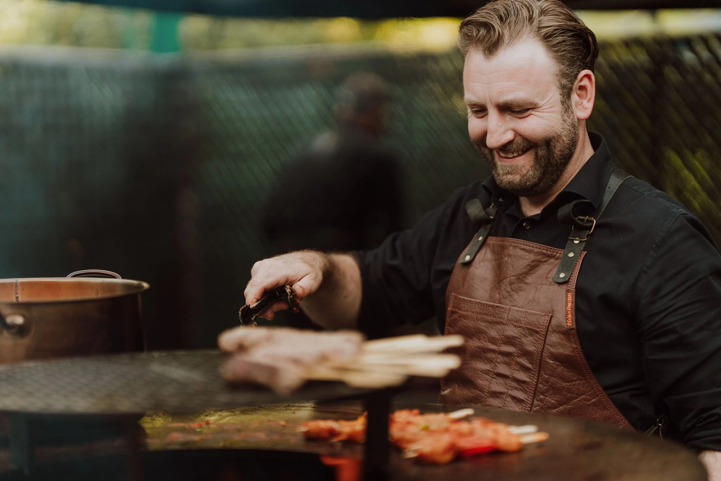 Man die buiten op een grill vlees bereidt, lachend terwijl hij tangen vasthoudt.